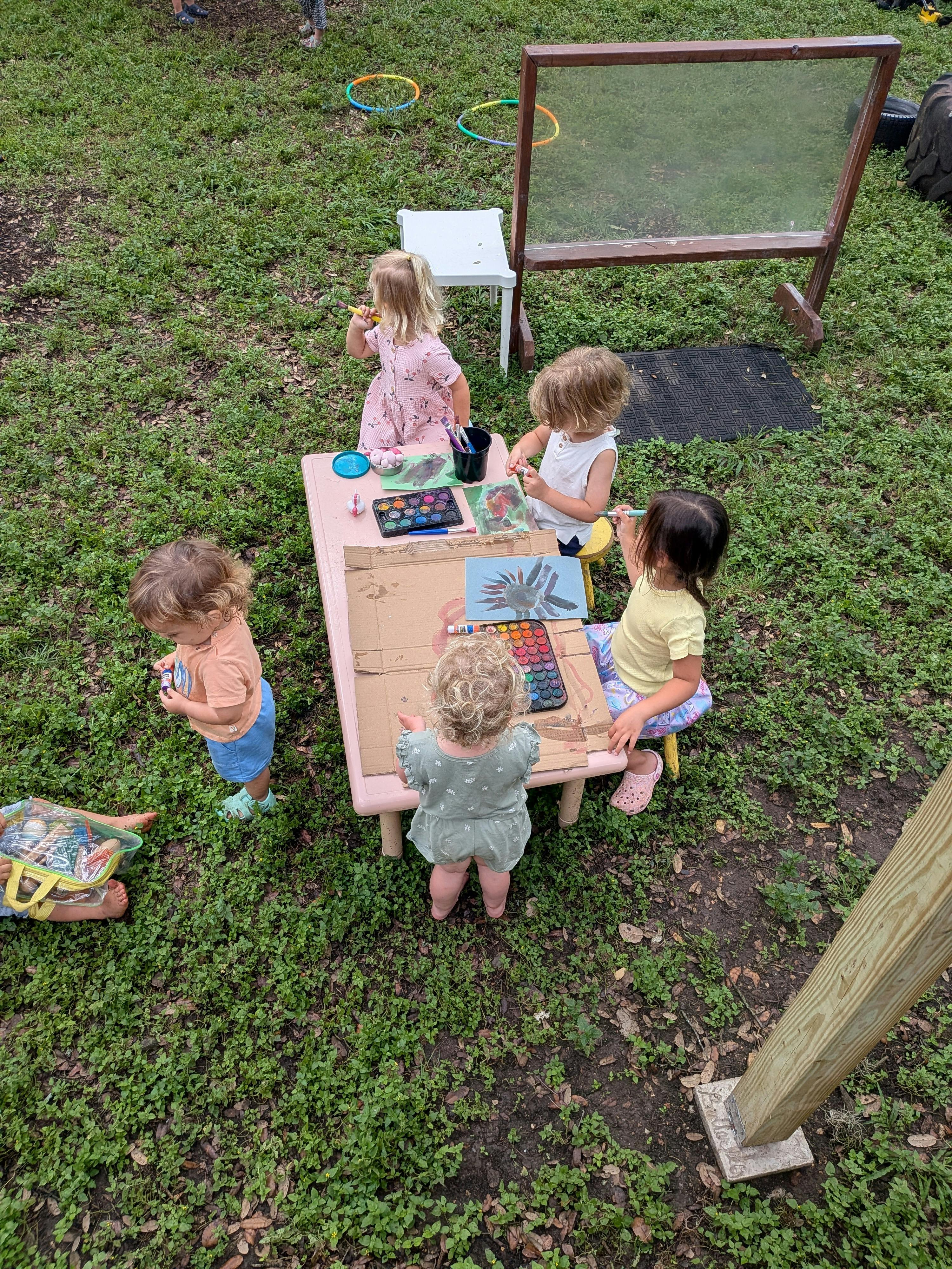 Several children painting and drawing at a small table outdoors