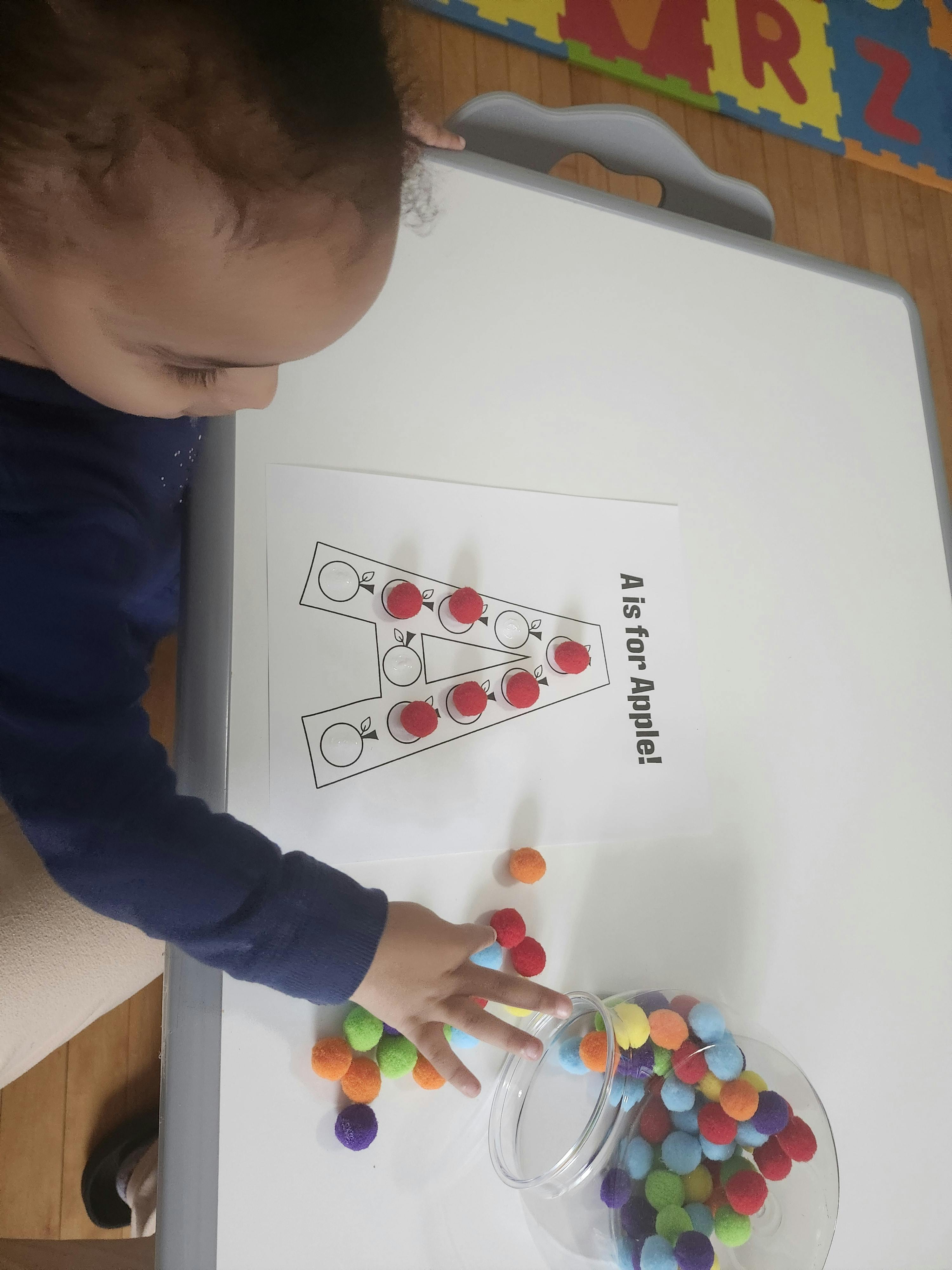 Child placing red pom-poms on an "A is for Apple" activity sheet