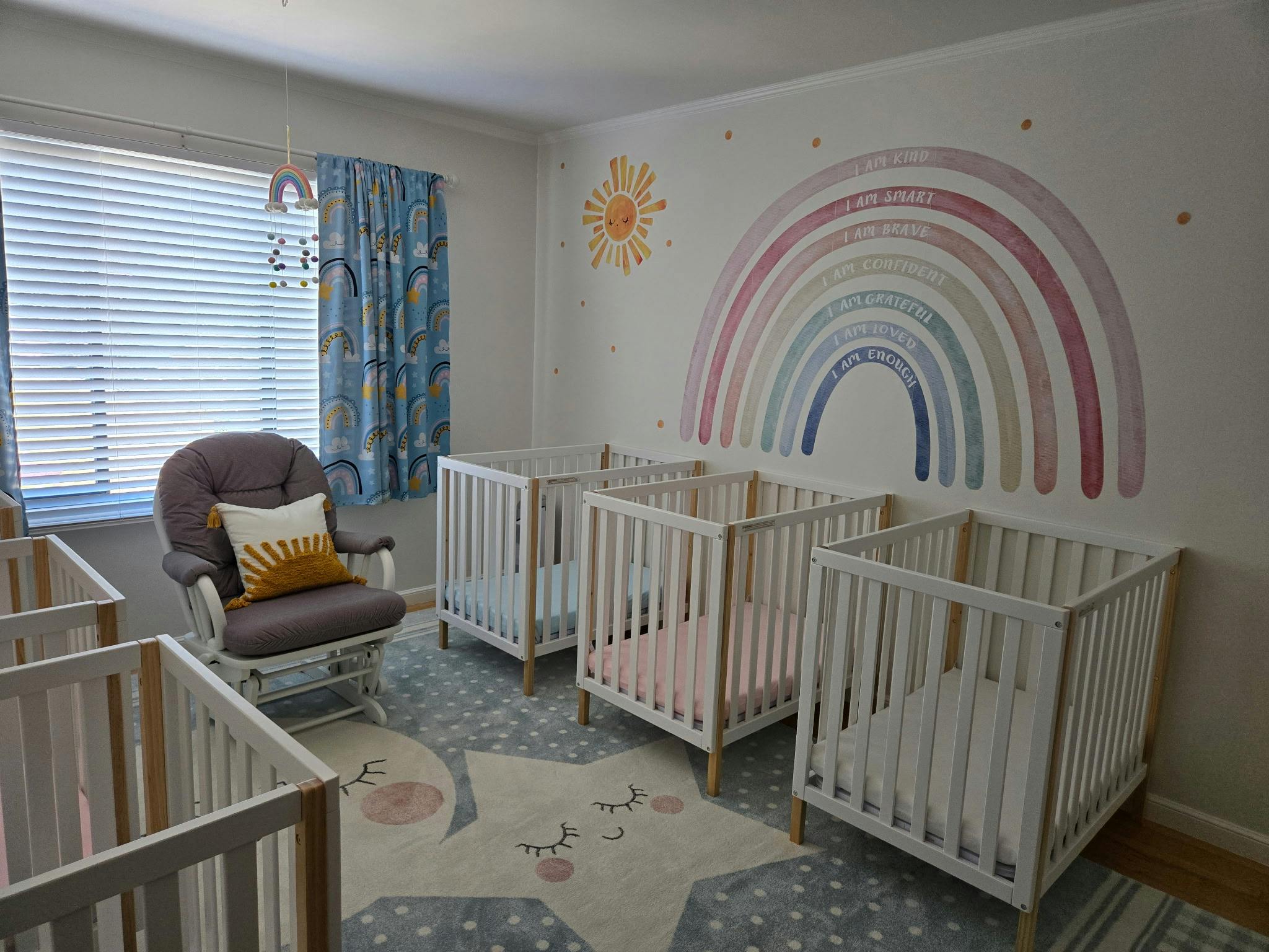 Room with multiple cribs, a rocking chair, and a rainbow wall decal