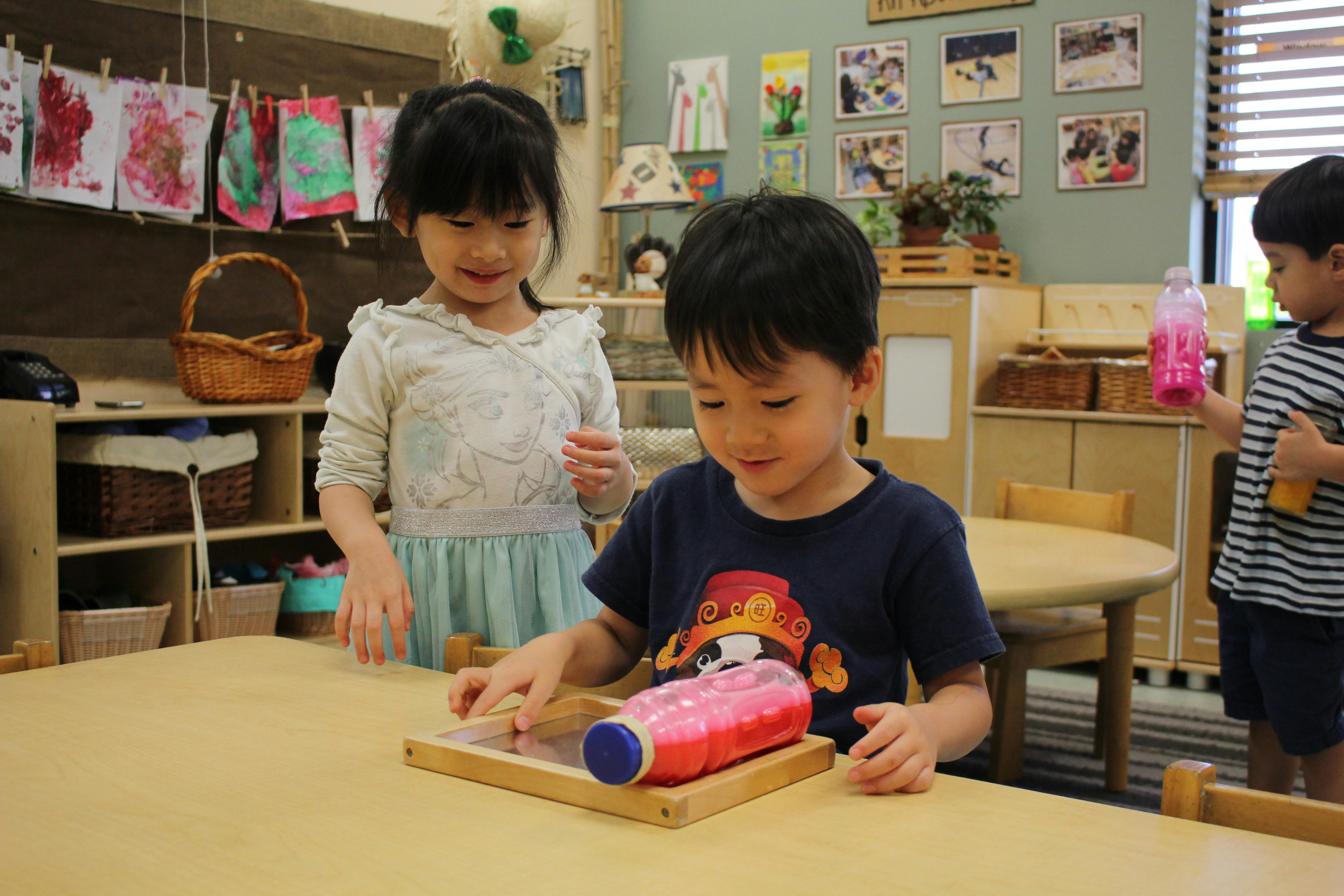 Child playing with a toy on a table in a classroom, with another child watching