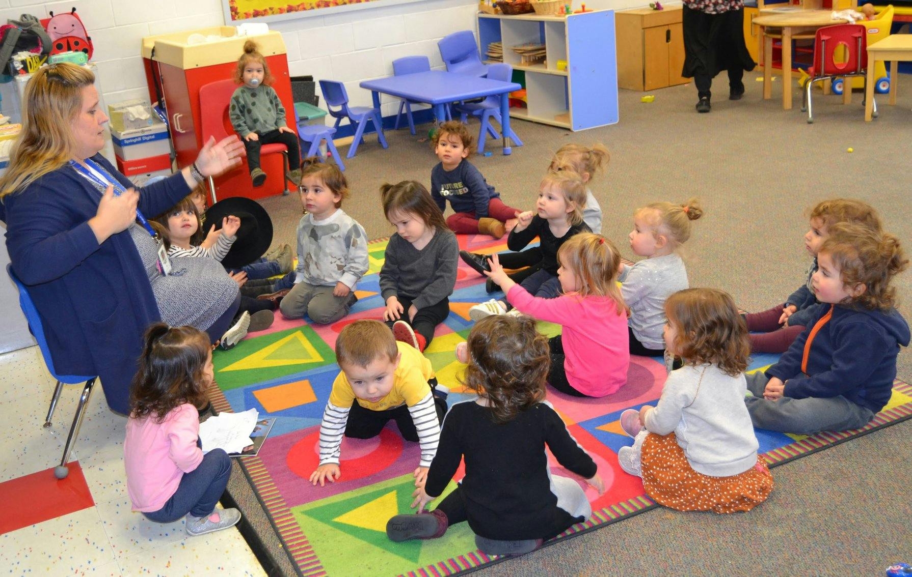 Adult speaking to a group of children sitting on a colorful rug in a classroom