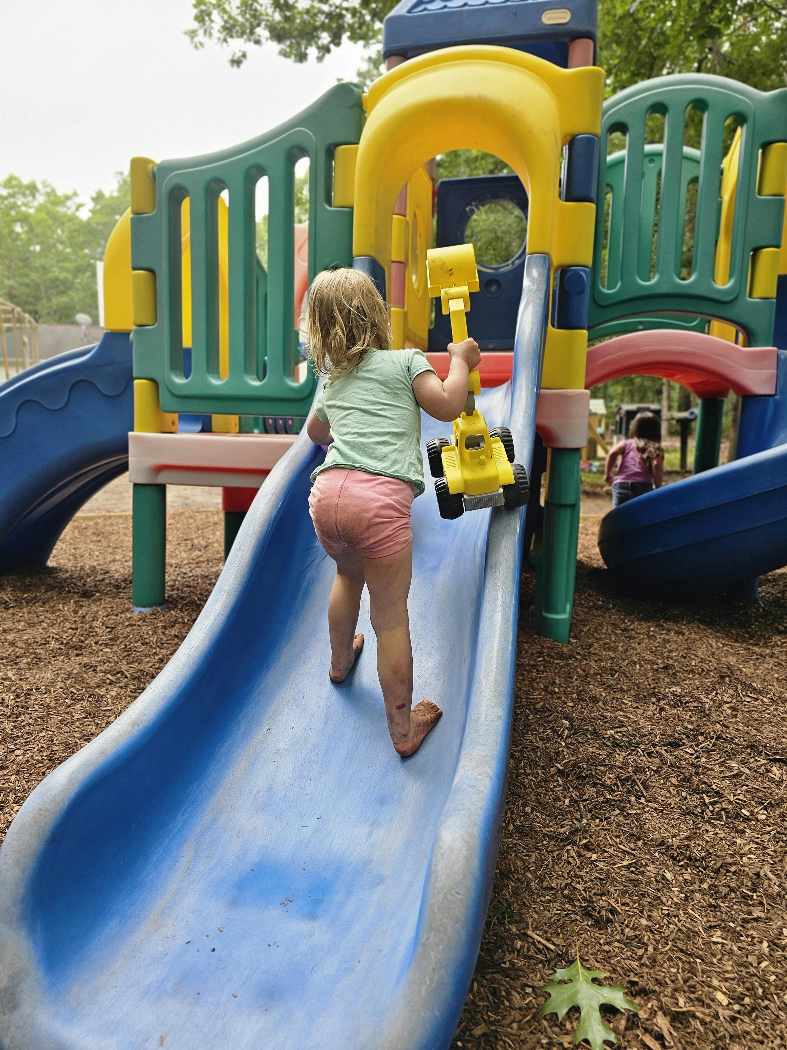 Child climbing up a blue slide on a playground while holding a toy