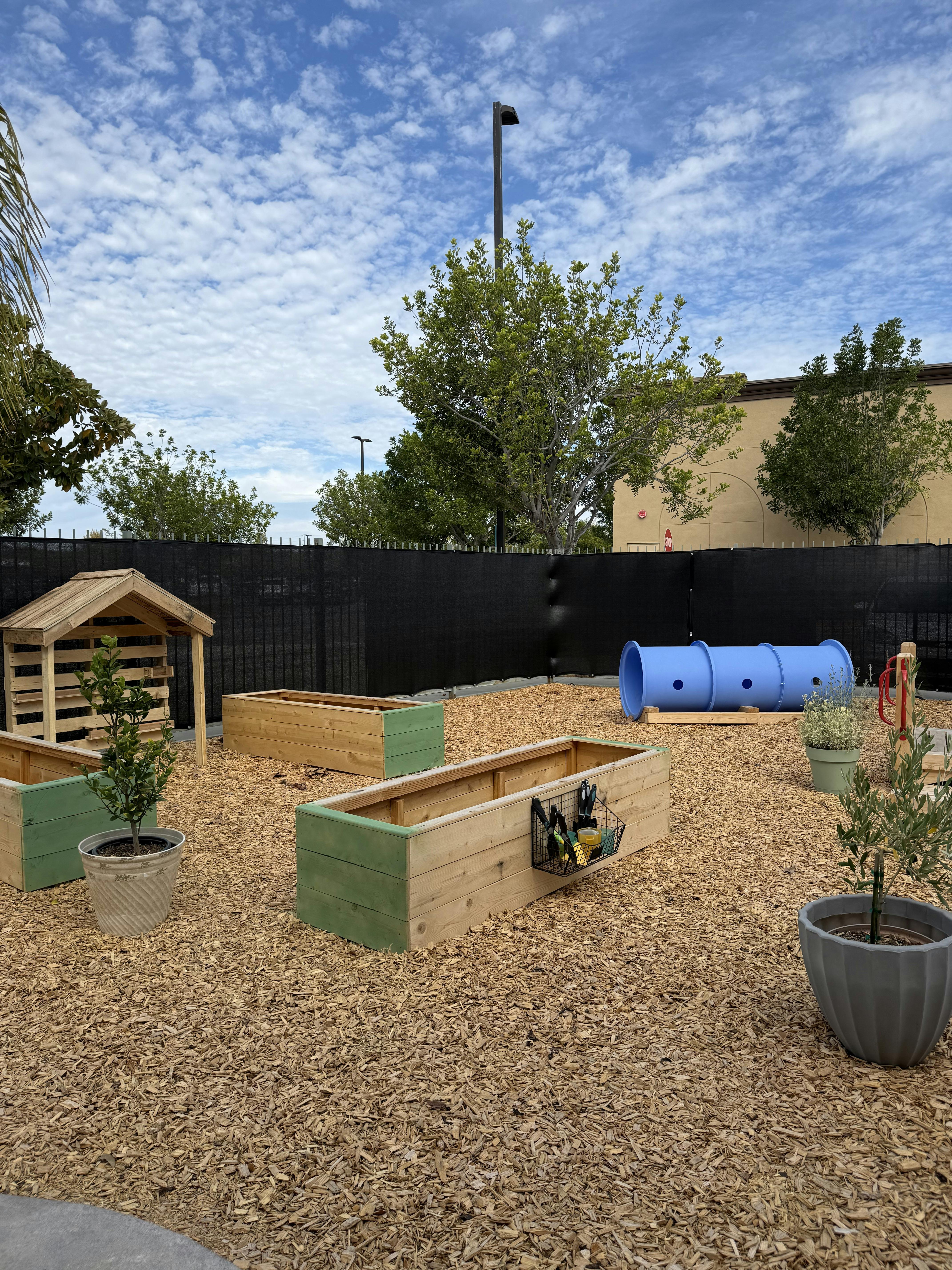 Outdoor play area with raised garden beds, a small wooden house, and a blue tunnel