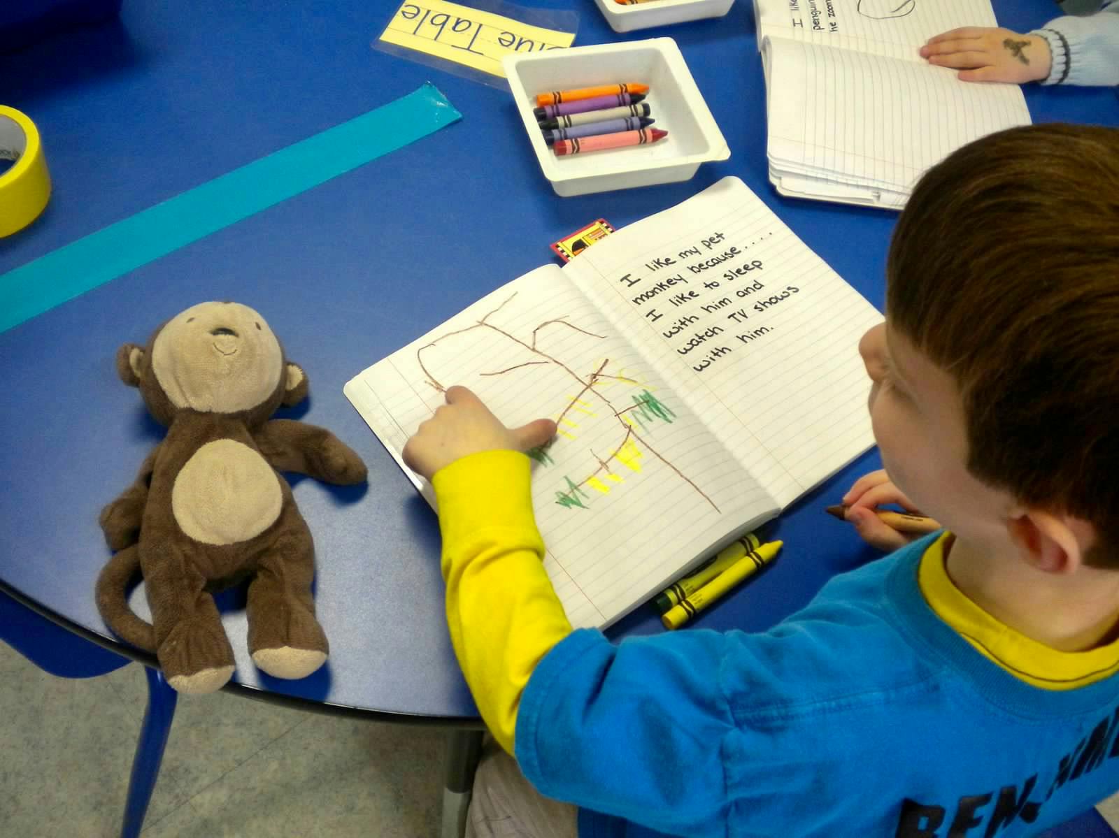 Child drawing in a notebook with a stuffed monkey on a blue table