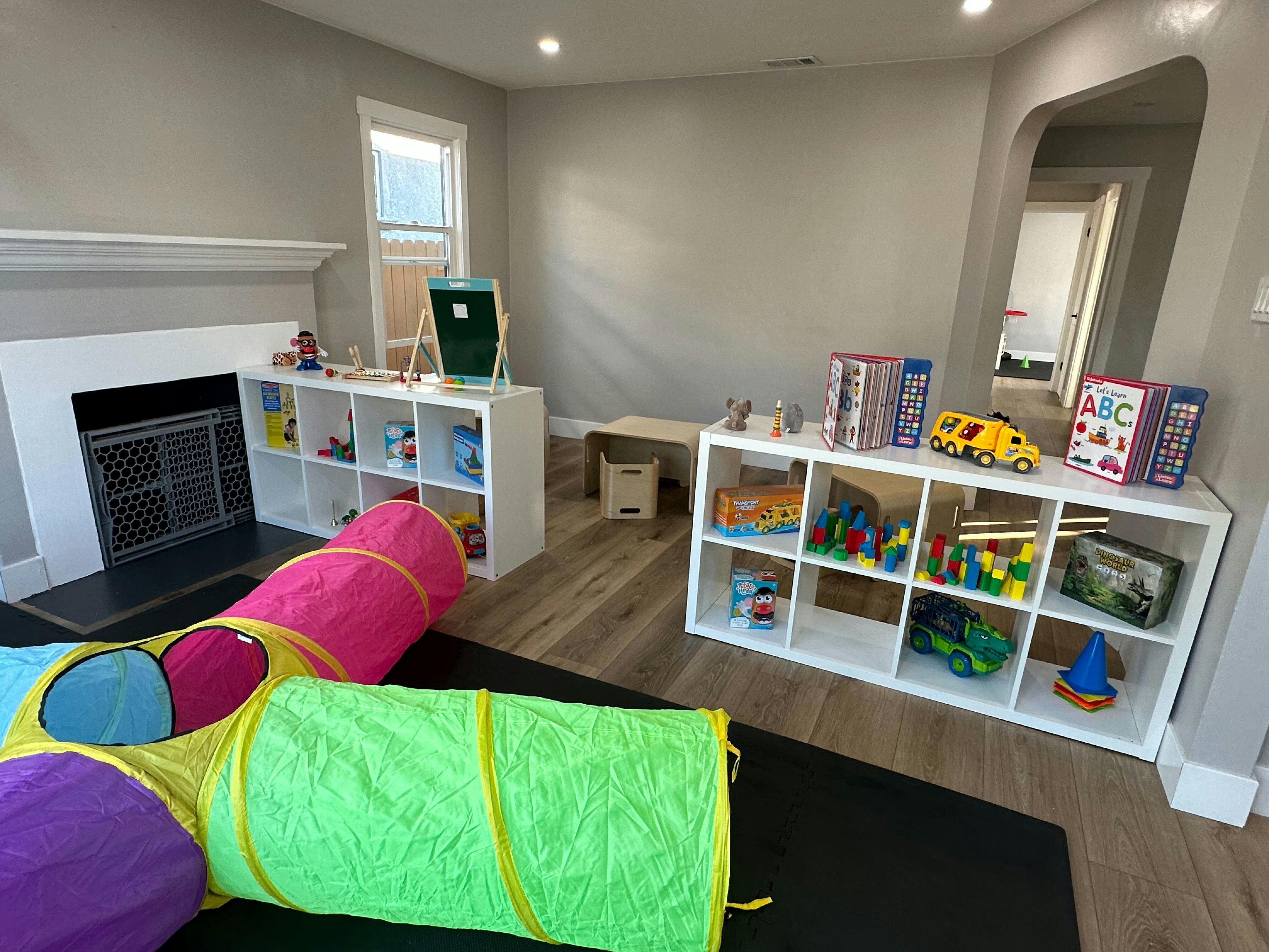 Playroom with colorful play tunnel, shelves of toys, and a small table