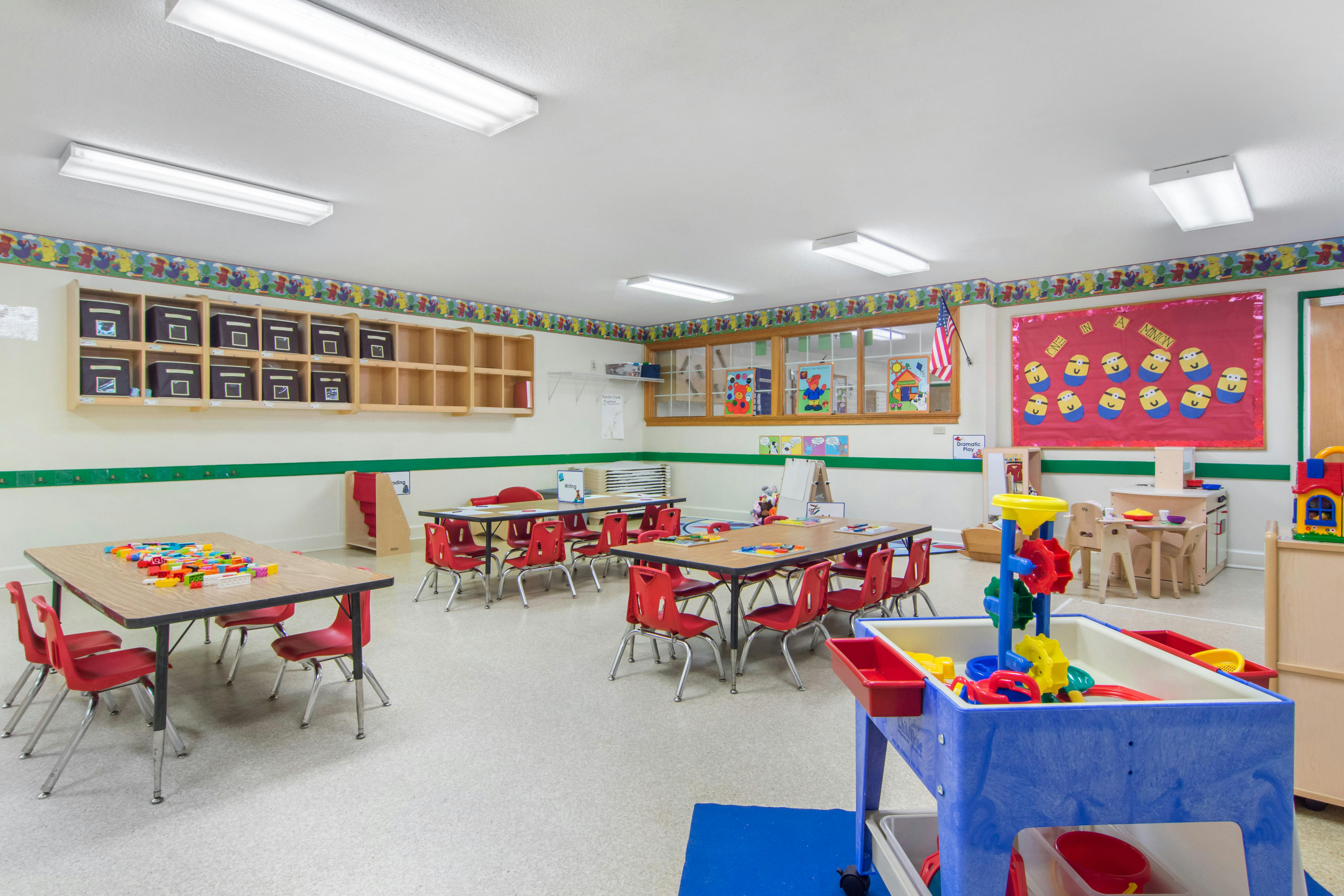 An empty early childhood classroom with multiple tables and chairs, shelves, and play areas