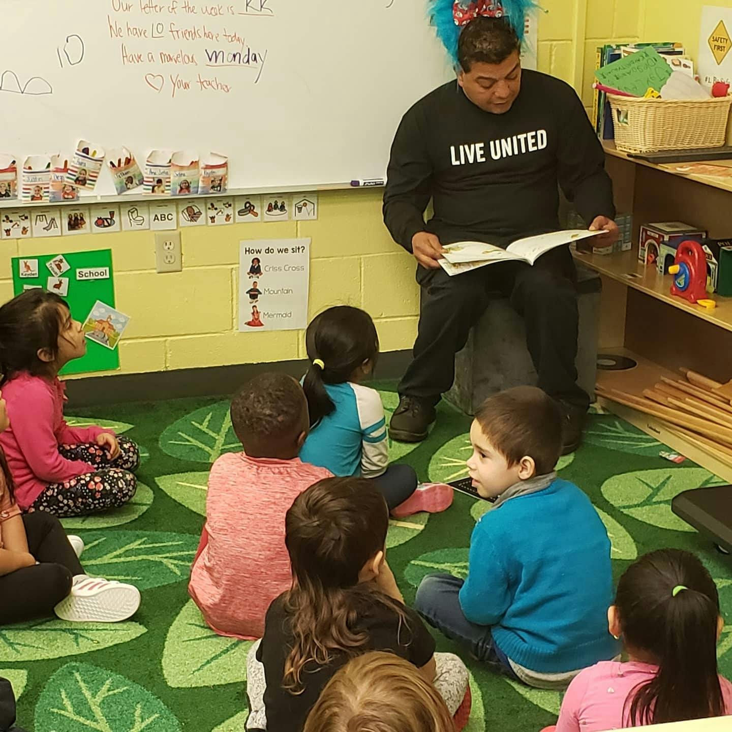 Person reading a book to a group of children sitting on a green rug