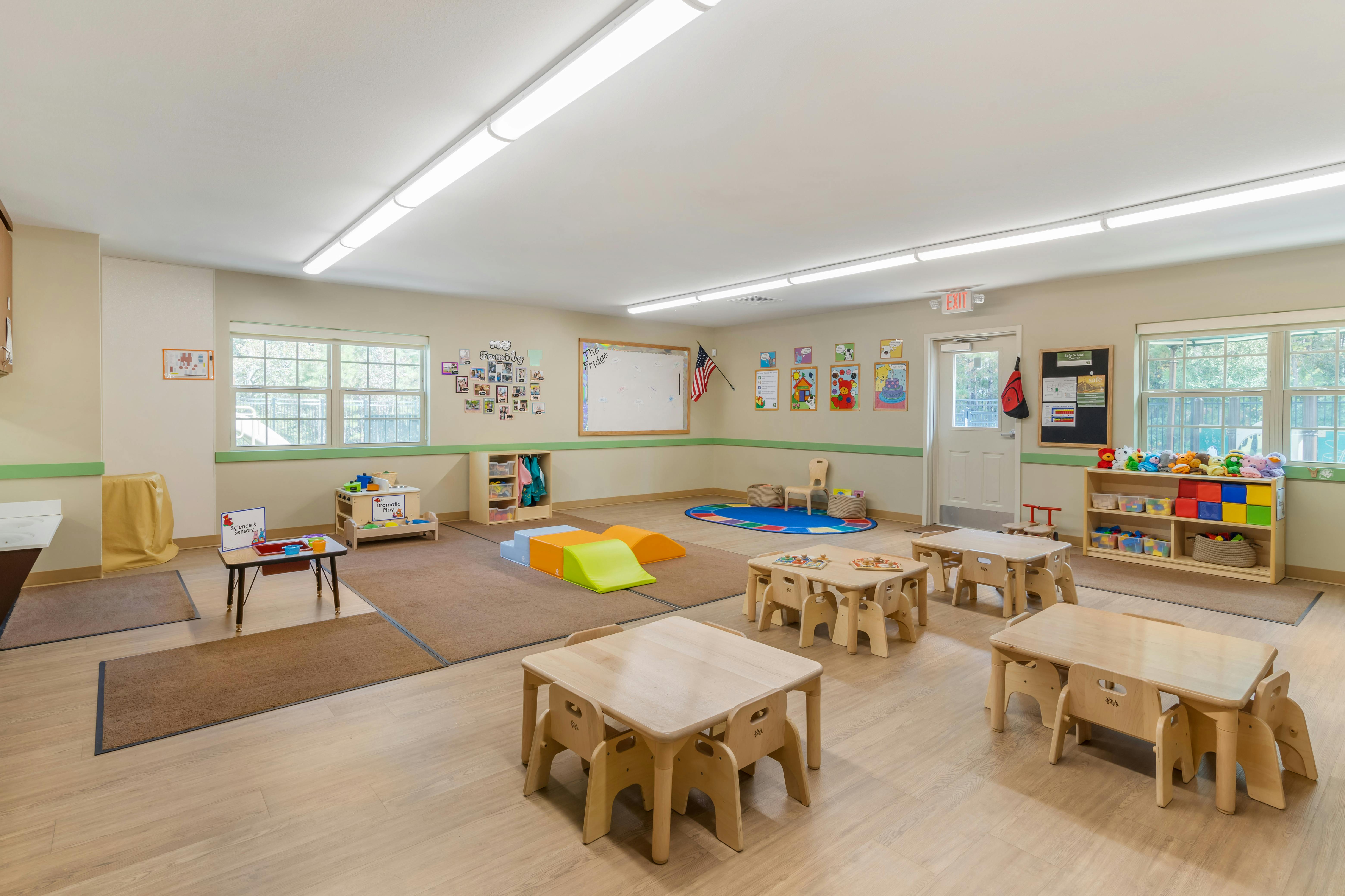 Empty classroom with small wooden tables and chairs, play mats, and toys