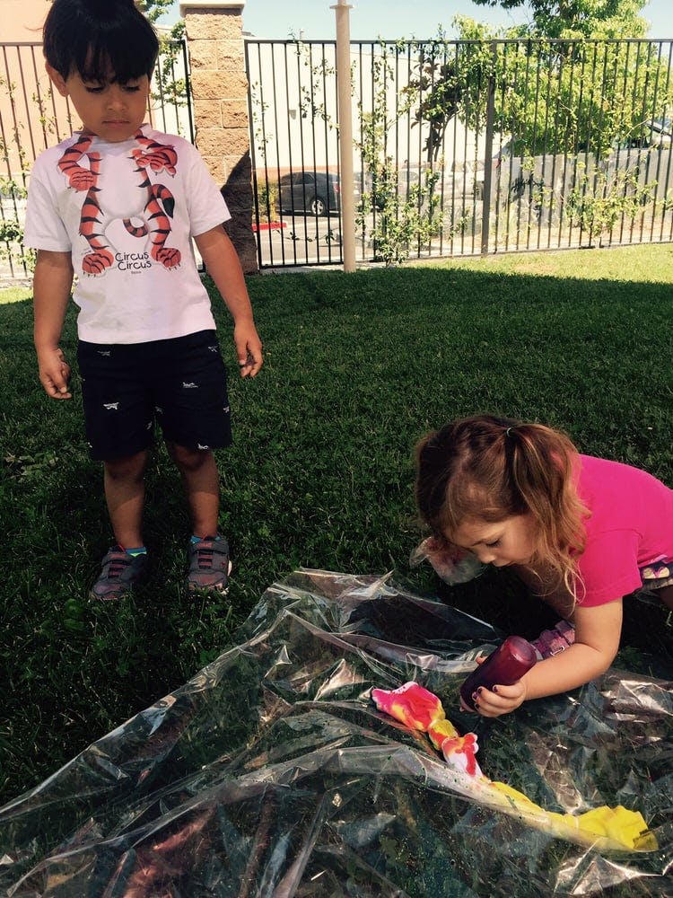 Child standing while another child paints on plastic on the grass
