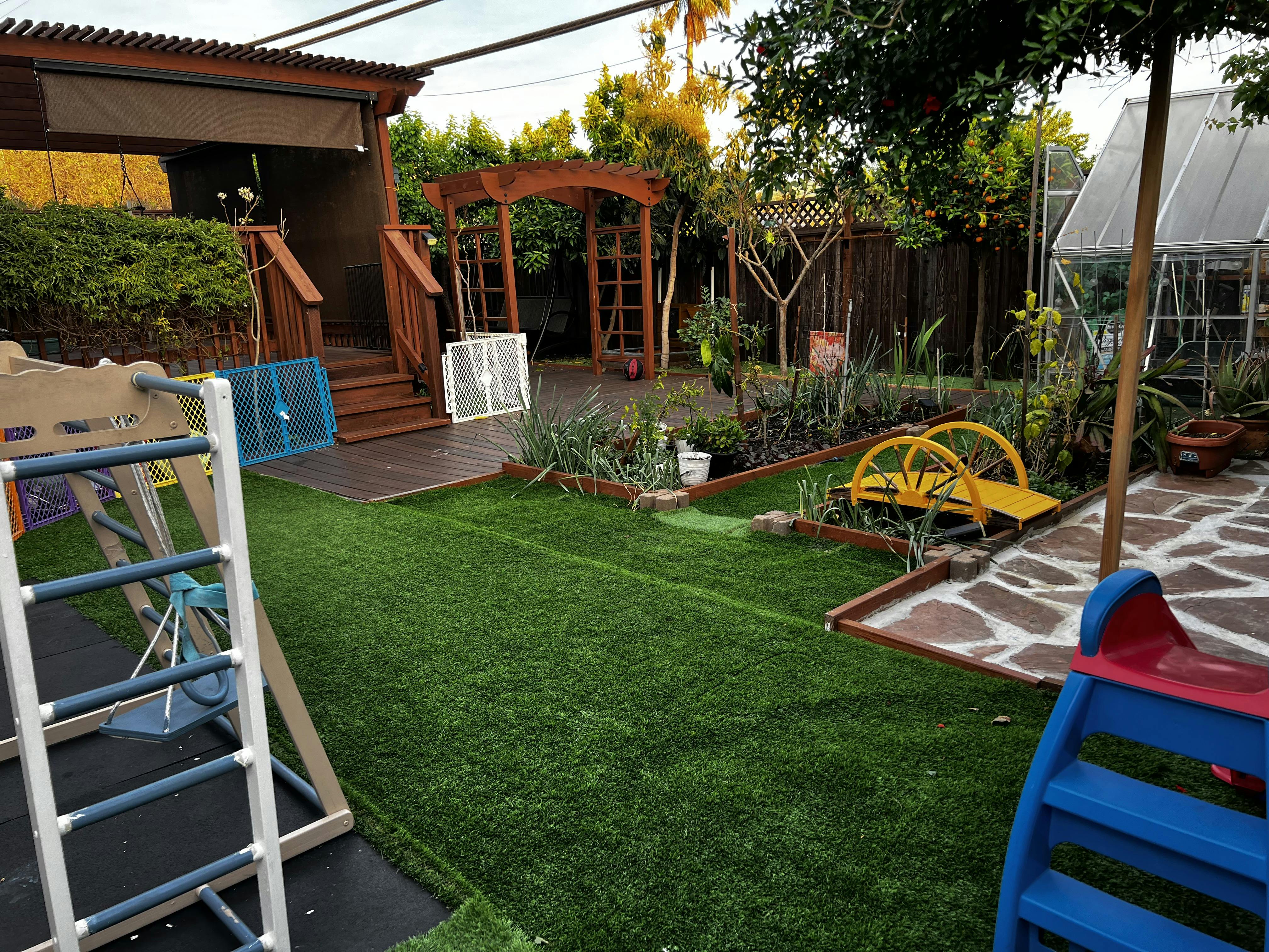 Backyard with artificial turf, play structures, wooden deck, and garden beds