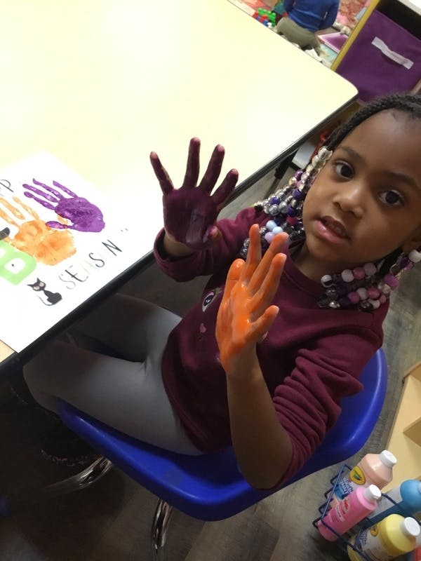 Child at a table with hands covered in paint, showing them to the camera