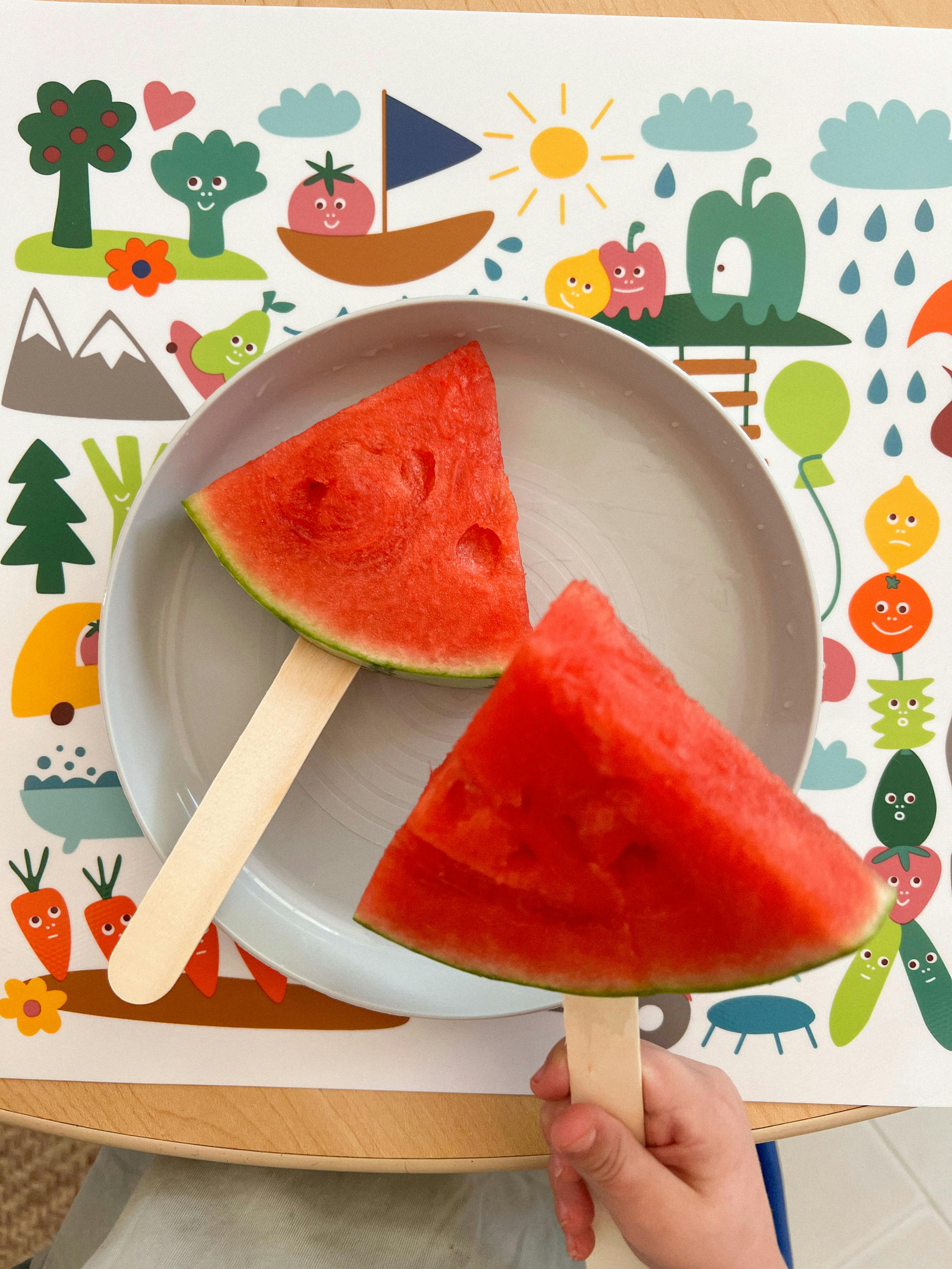 Two watermelon slices on sticks, one held by a child's hand