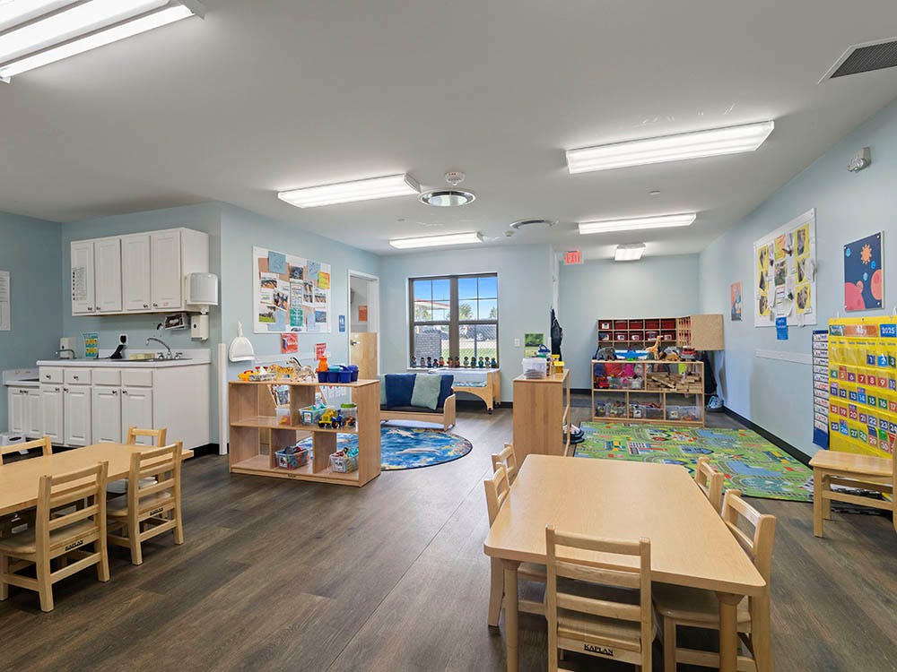 A brightly lit classroom with small tables and chairs, a play area, and a kitchen counter