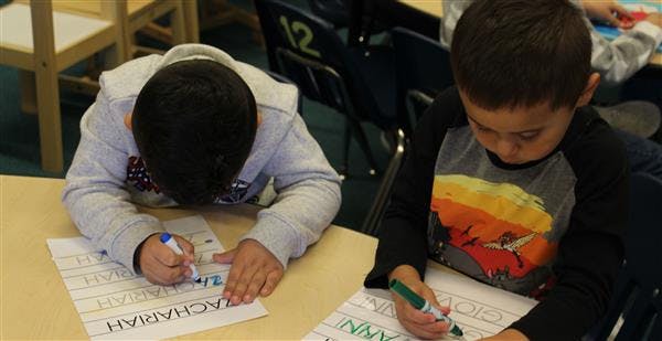 Two children writing on paper at a desk in a classroom