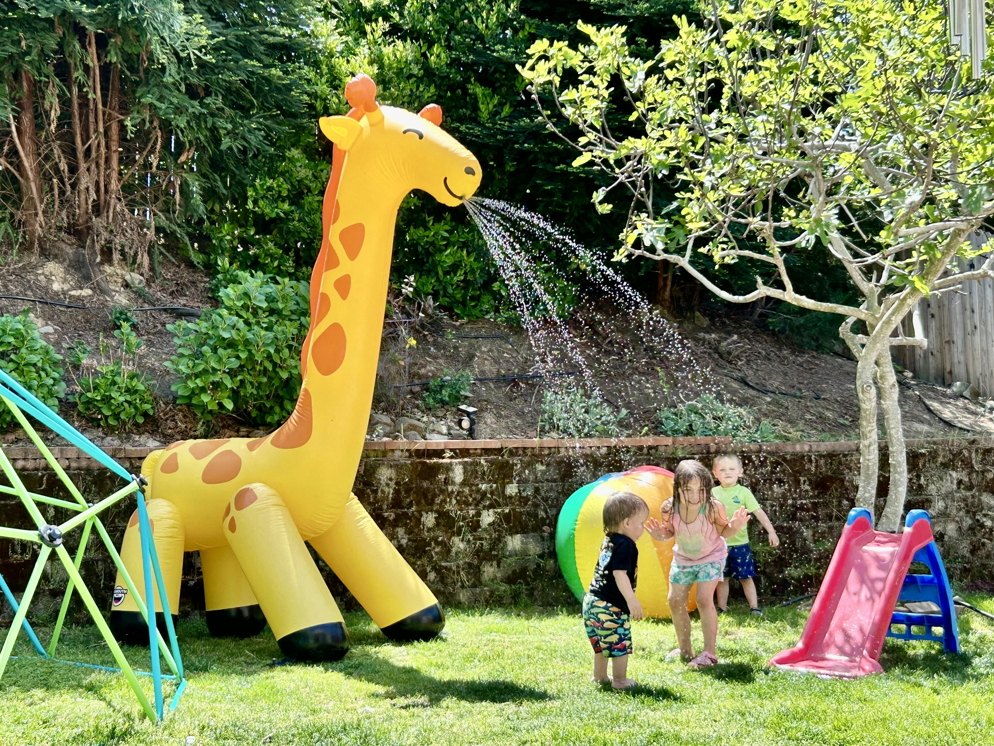 Inflatable giraffe sprinkler spraying water over three children in a backyard