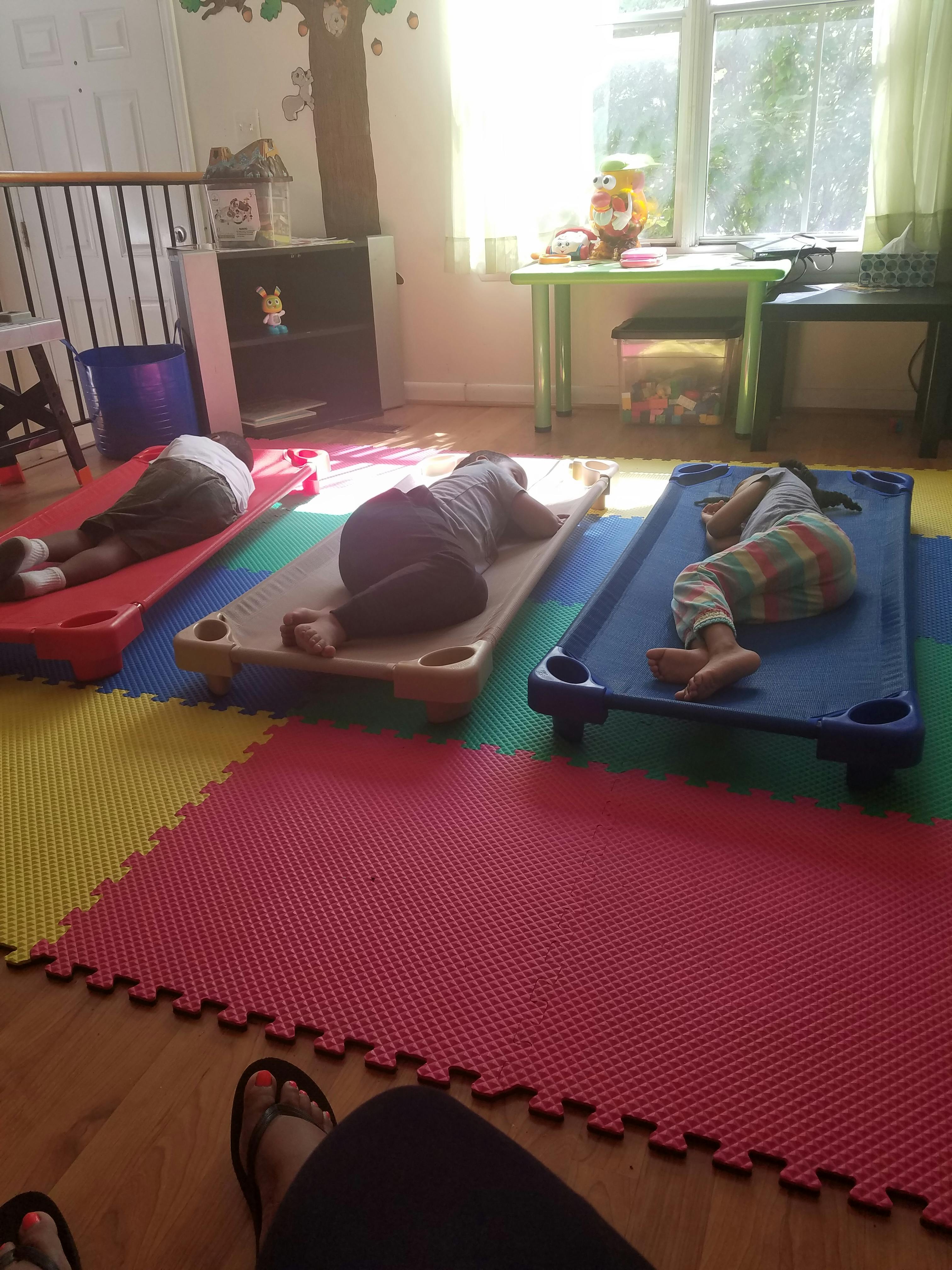 Three children resting on small cots on colorful mats in a playroom