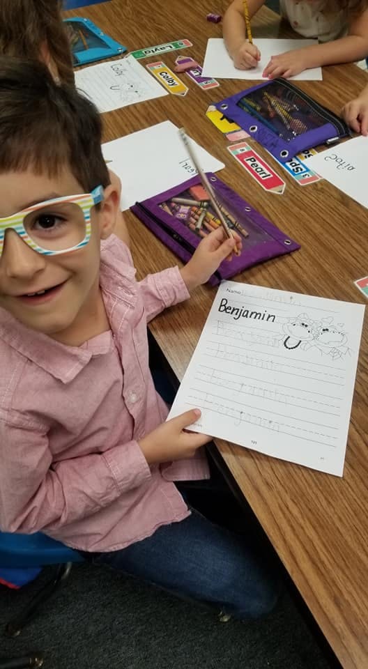 Child in glasses holding a pencil and a paper with "Benjamin" written on it