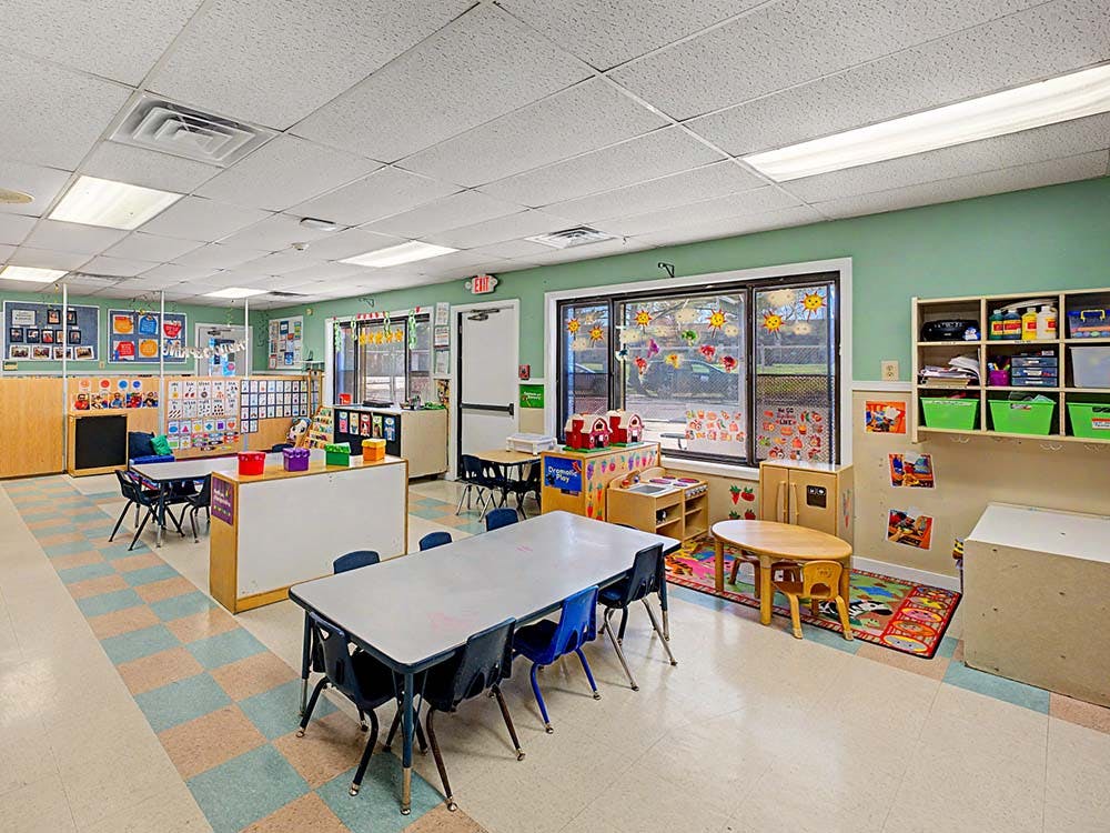 Classroom with multiple tables and chairs, shelves with toys, and window decals