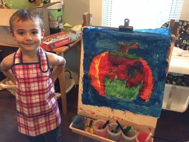 Child in apron standing next to an easel with a painting