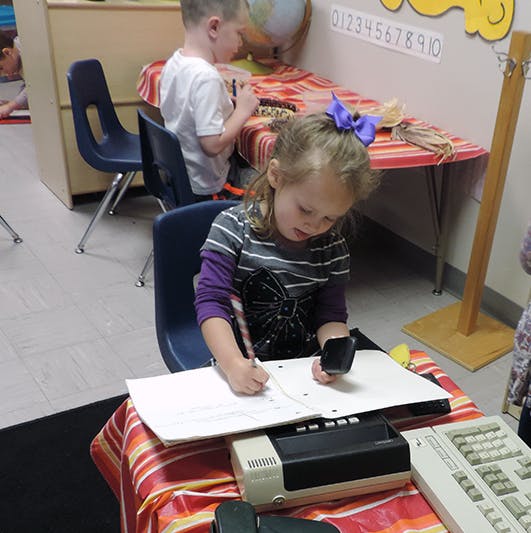 Child writing at a desk with another child in the background