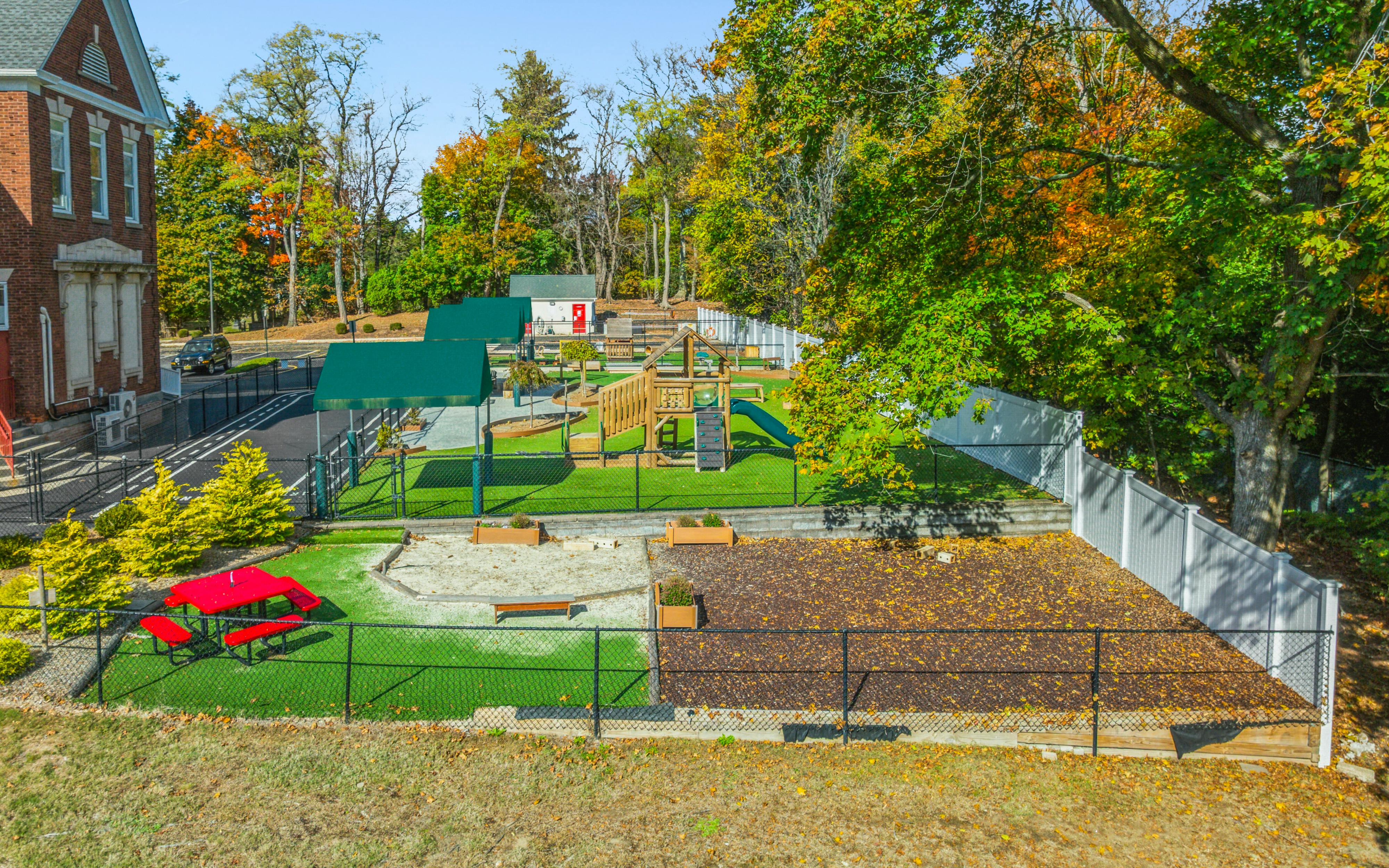 Outdoor playground with sandpit, wooden play structure, red picnic table, and artificial grass