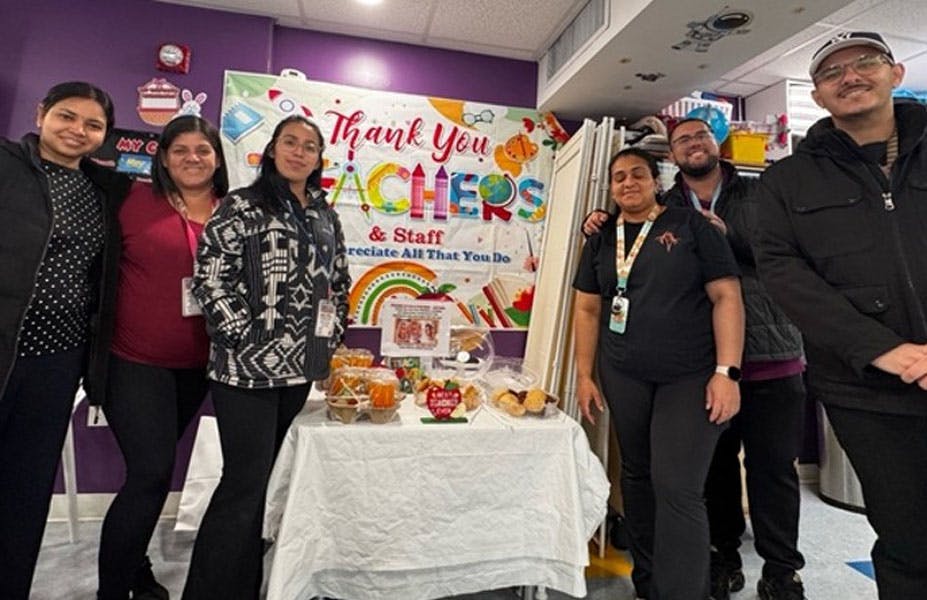 Group of people standing behind a table with food in front of a teacher sign