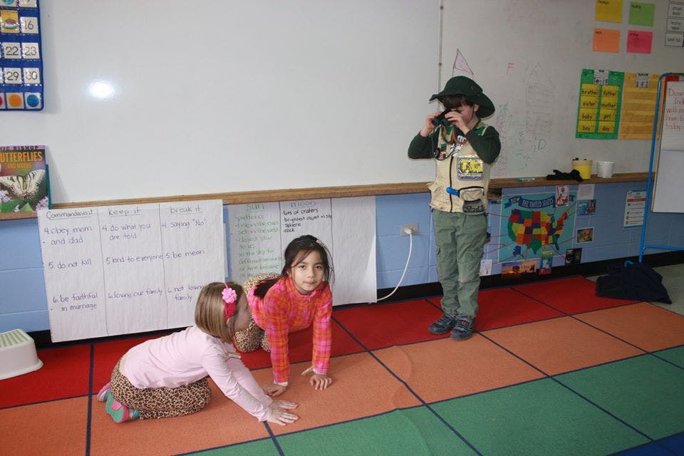 Three children playing in a classroom, one child in a safari hat holding binoculars