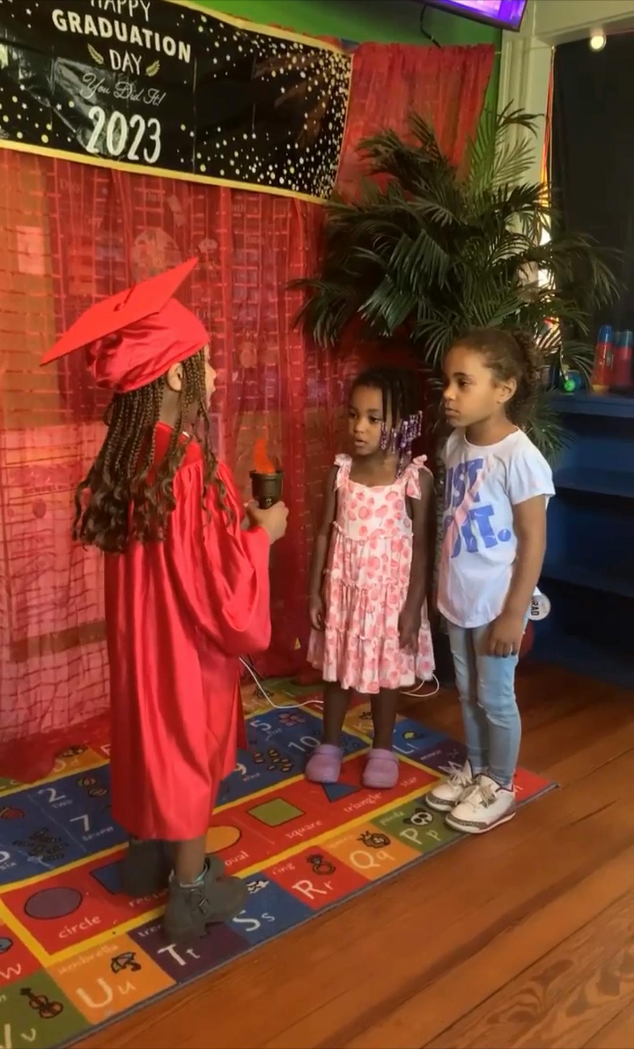 Child in graduation cap and gown holding a torch, facing two other children