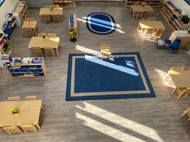 An empty classroom with small wooden tables and chairs, shelves, and blue rugs