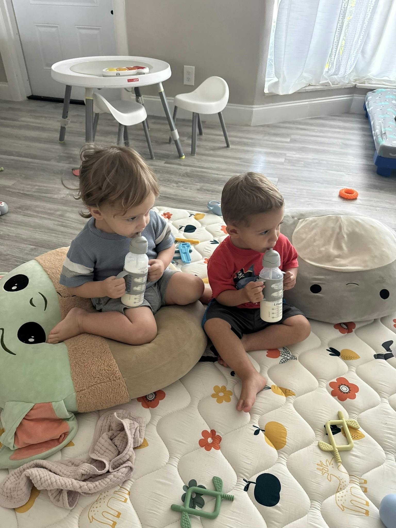 Two children sitting on a play mat holding baby bottles