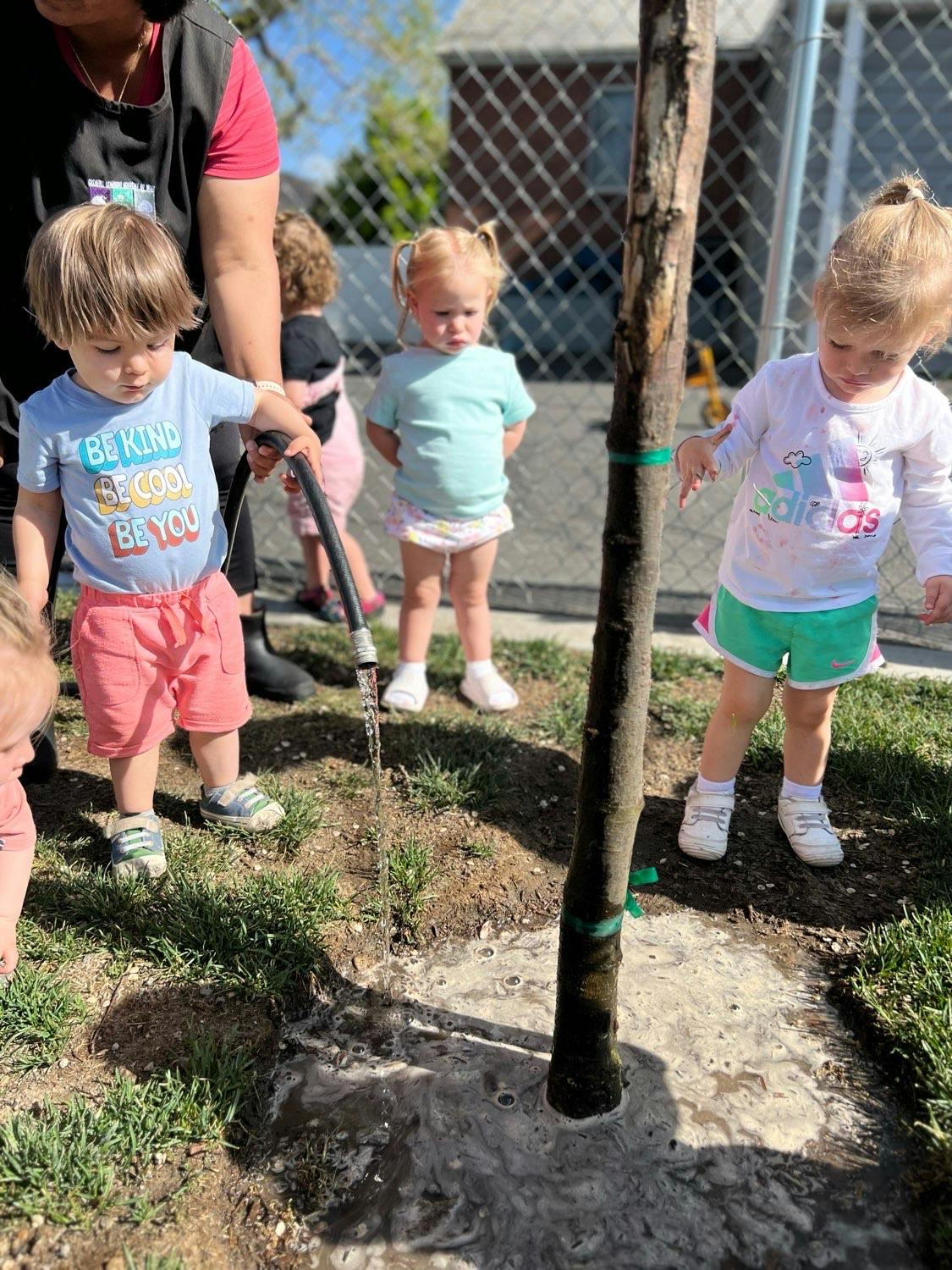 Child watering a newly planted tree with a hose, other children nearby