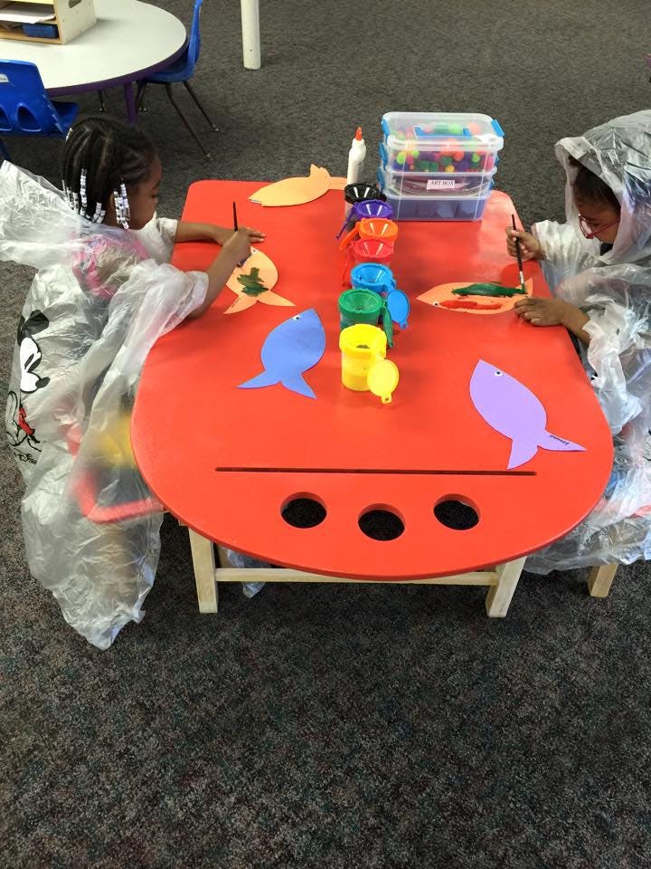 Two children painting fish cutouts at a red table in a classroom