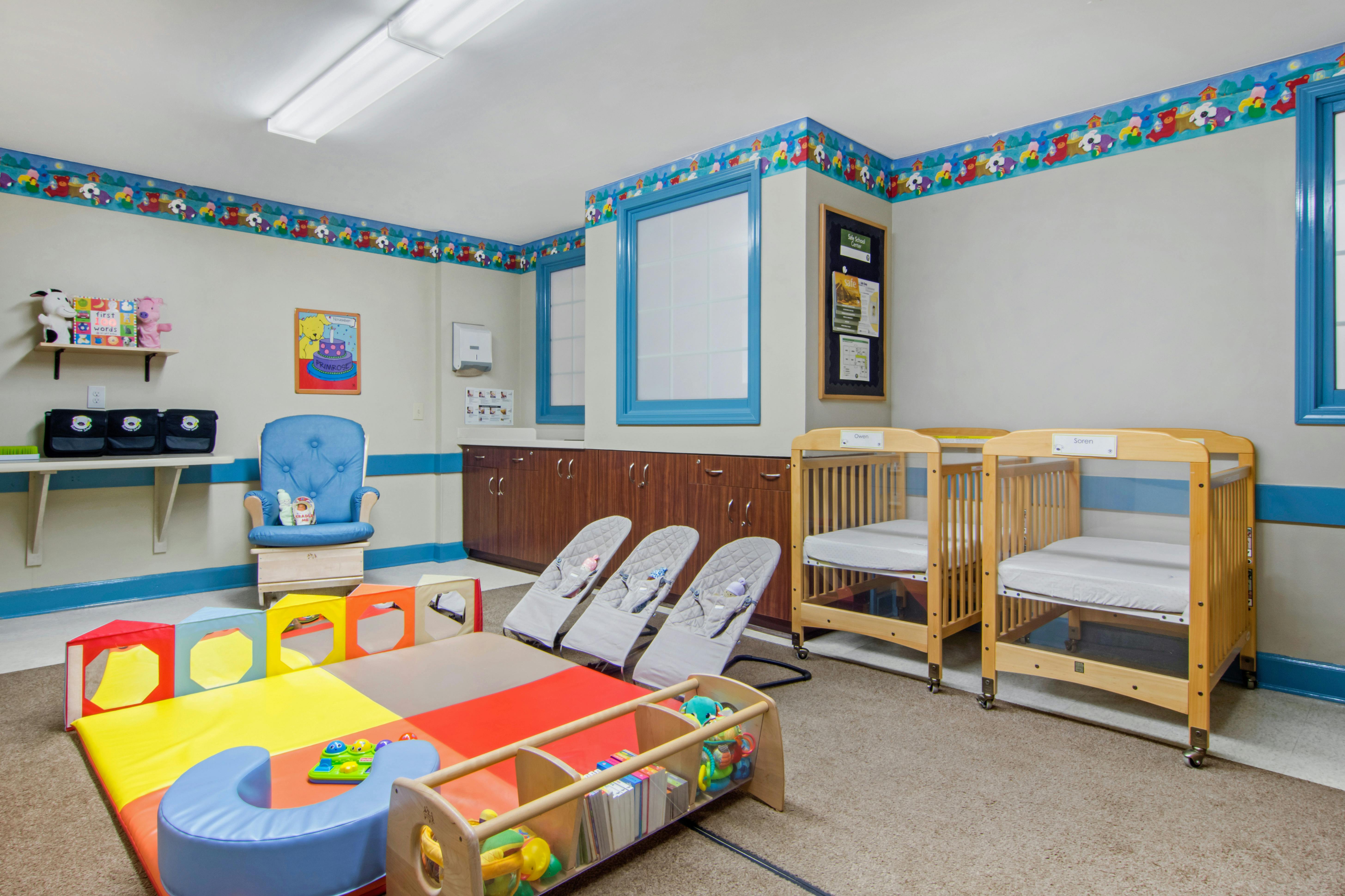 A colorful childcare room with cribs, bouncers, a playmat, and a blue armchair