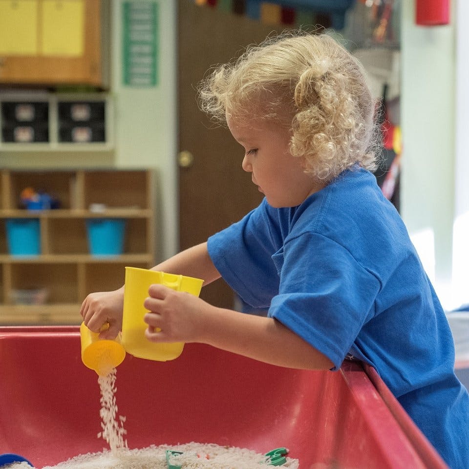 Child pouring white grains from a yellow cup into a red bin