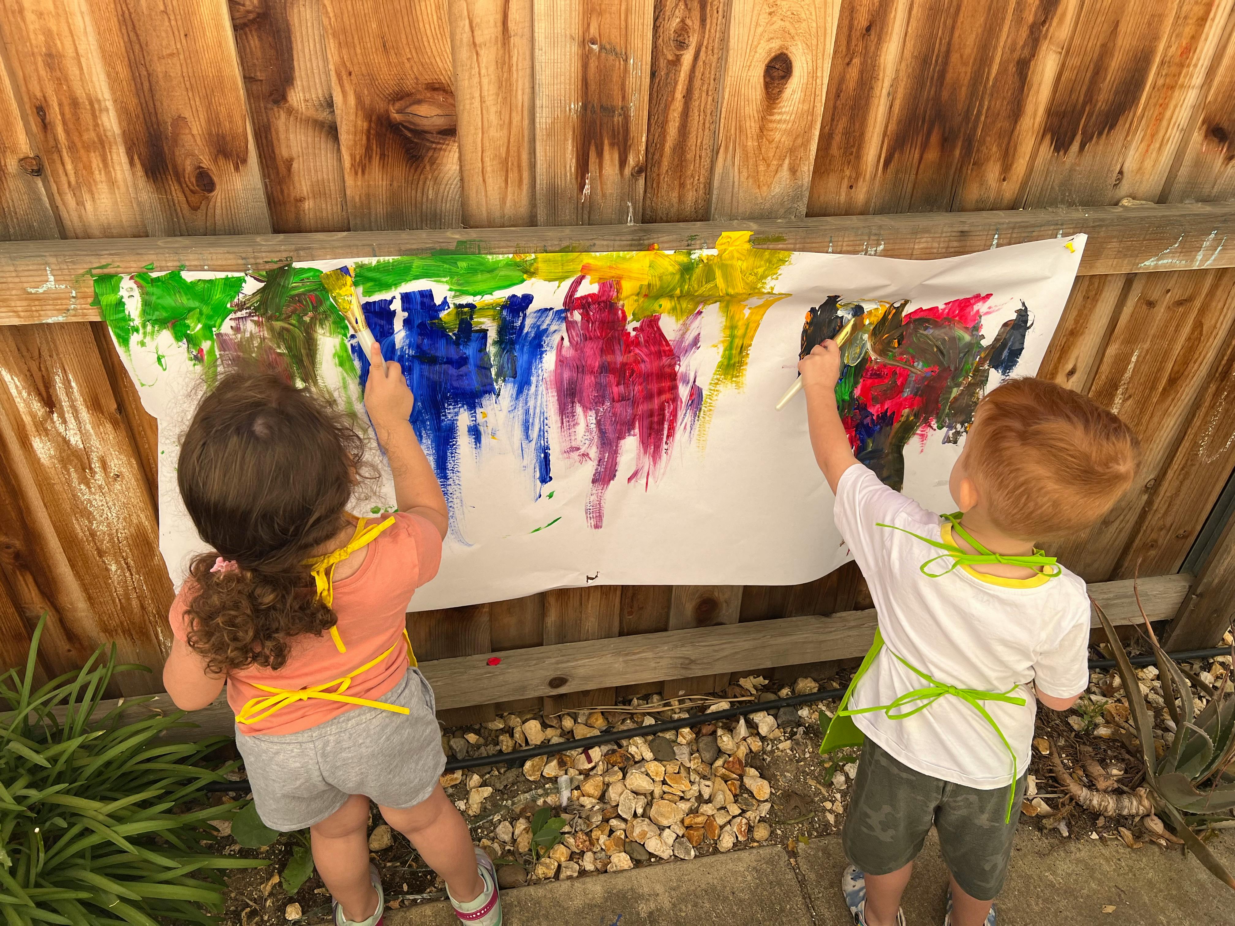 Two children painting on a large sheet of paper attached to a wooden fence