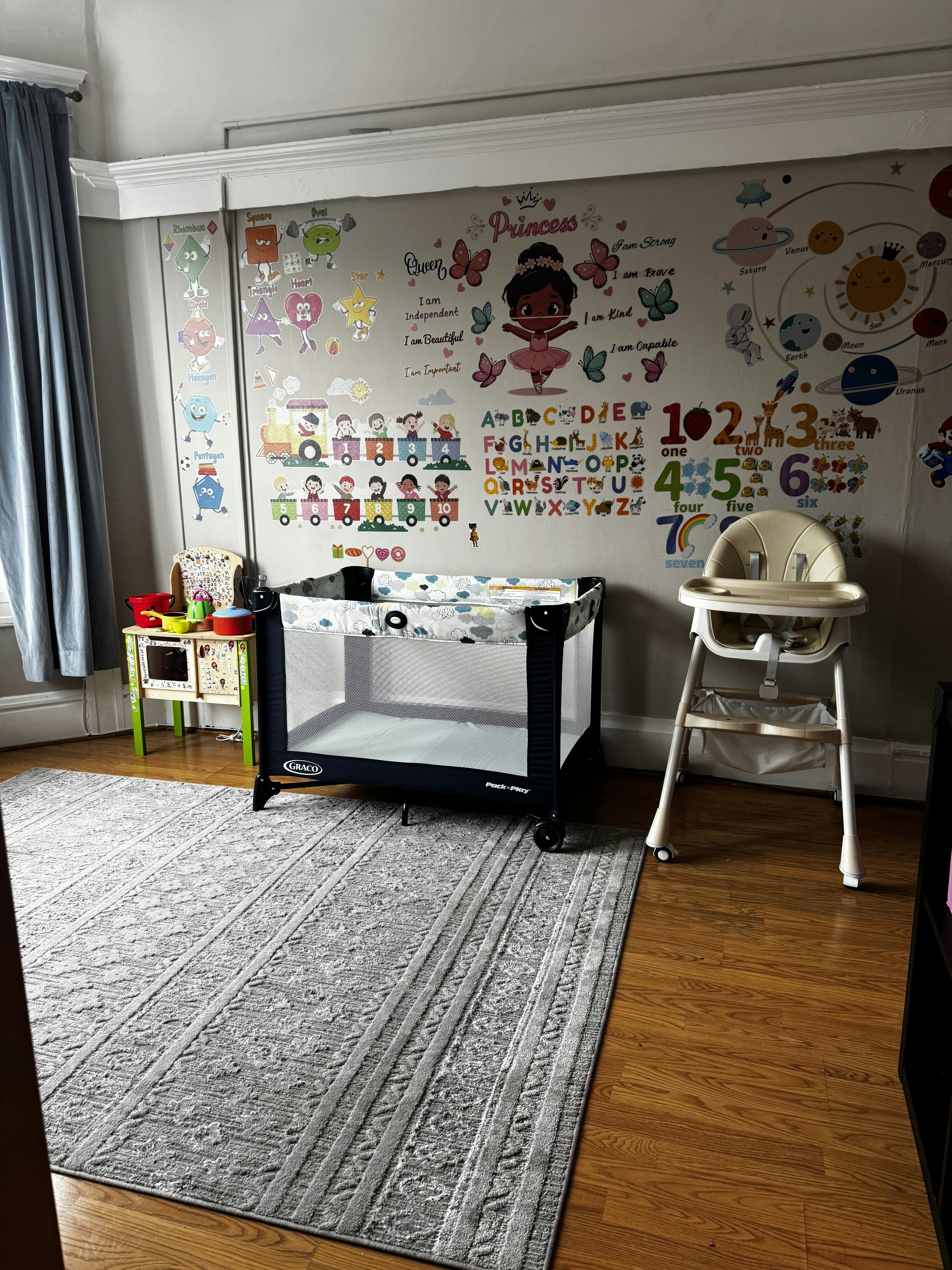A child's room with an educational mural, pack and play, and high chair