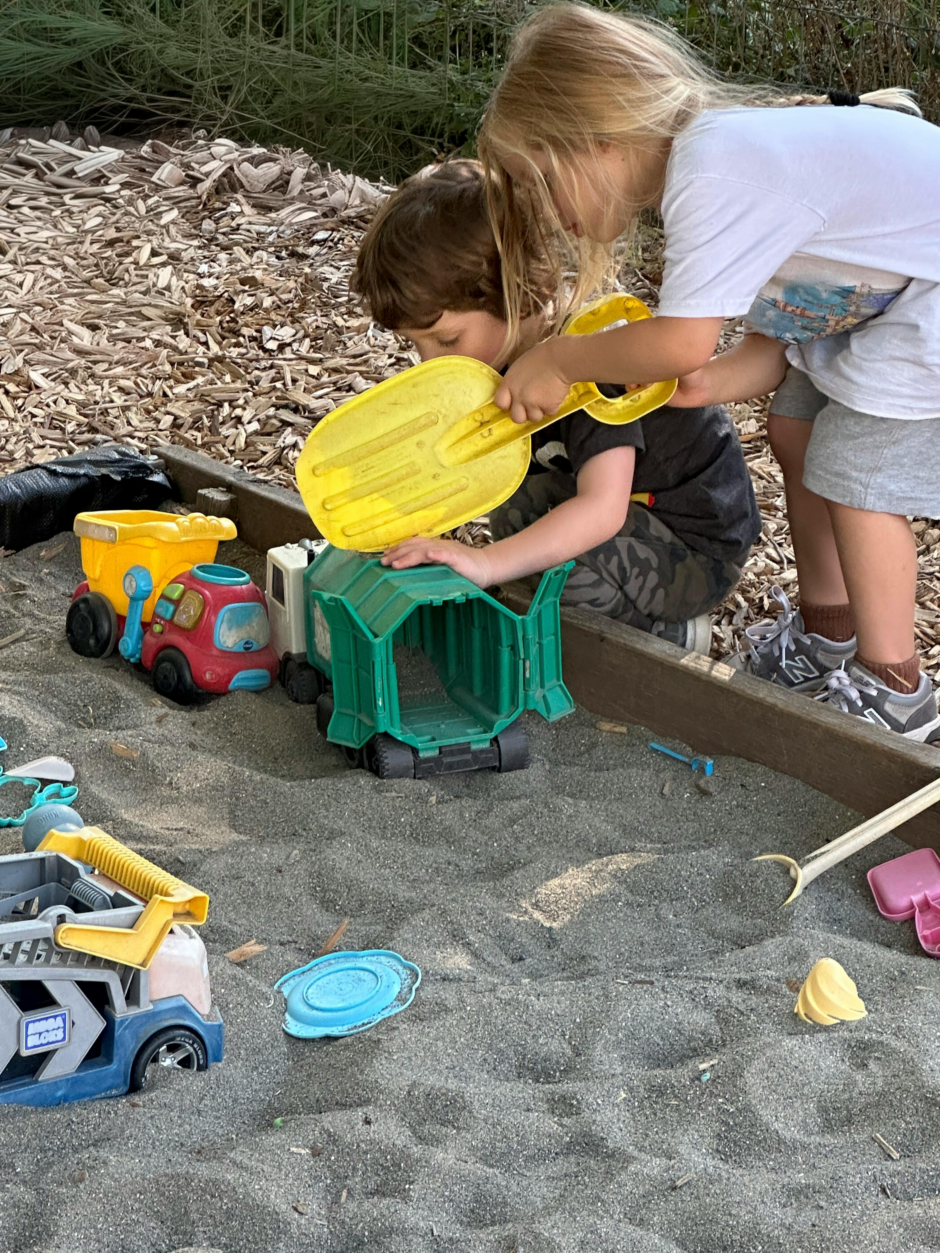 Two children playing with toy trucks and a shovel in a sandbox