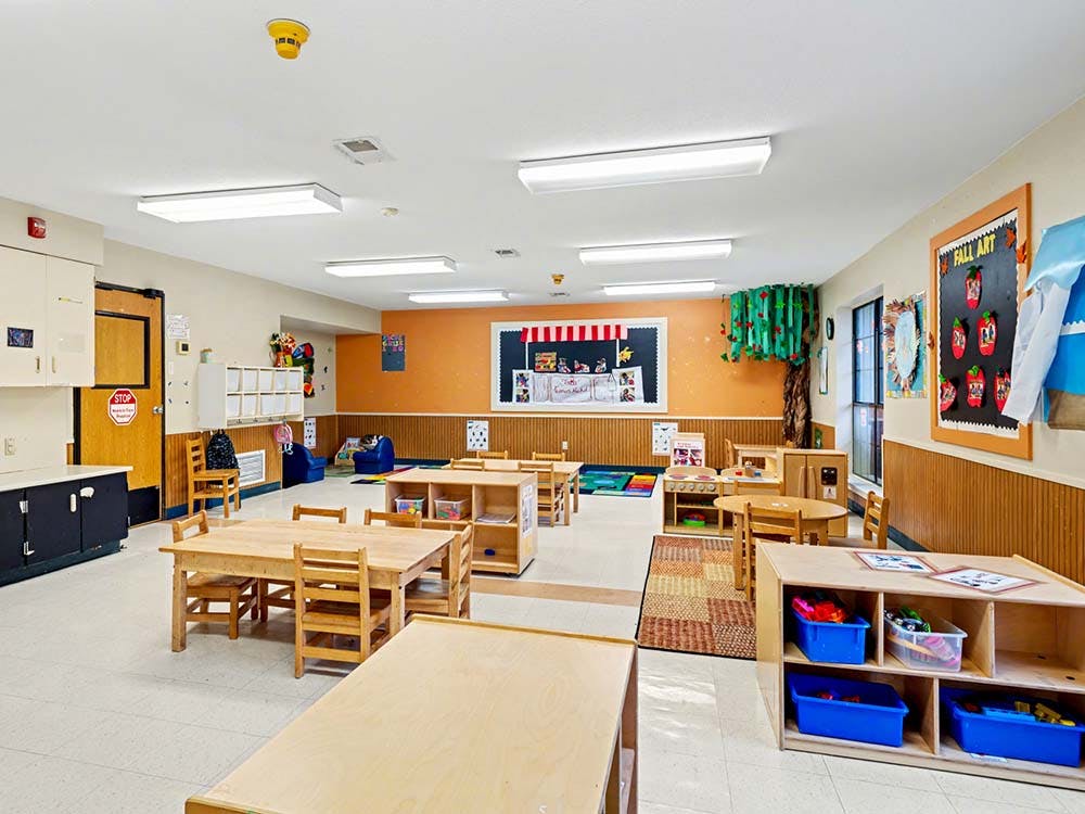 Empty classroom with small wooden tables and chairs, colorful rugs, and artwork