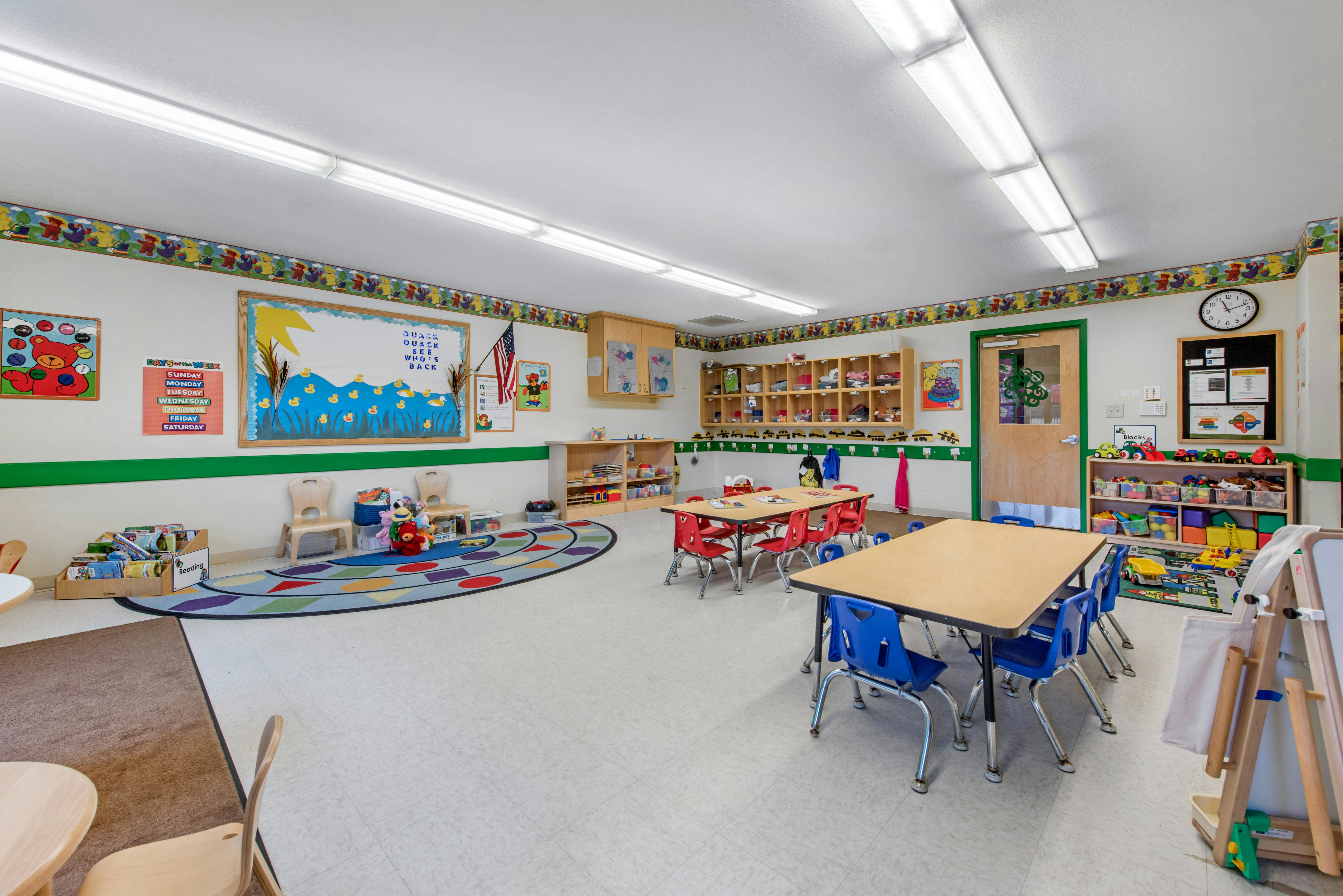 Classroom with tables, chairs, colorful rug, shelves, and educational decorations