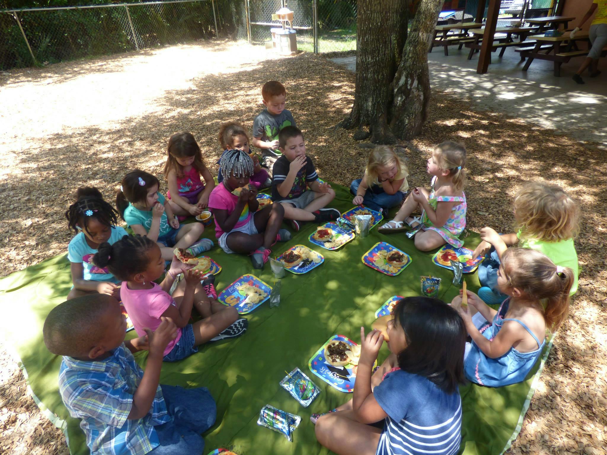 Children sitting on a green blanket eating snacks on the ground eating