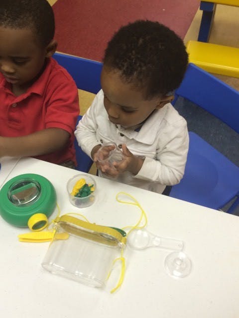 Child holding a clear container at a table with other children and toys