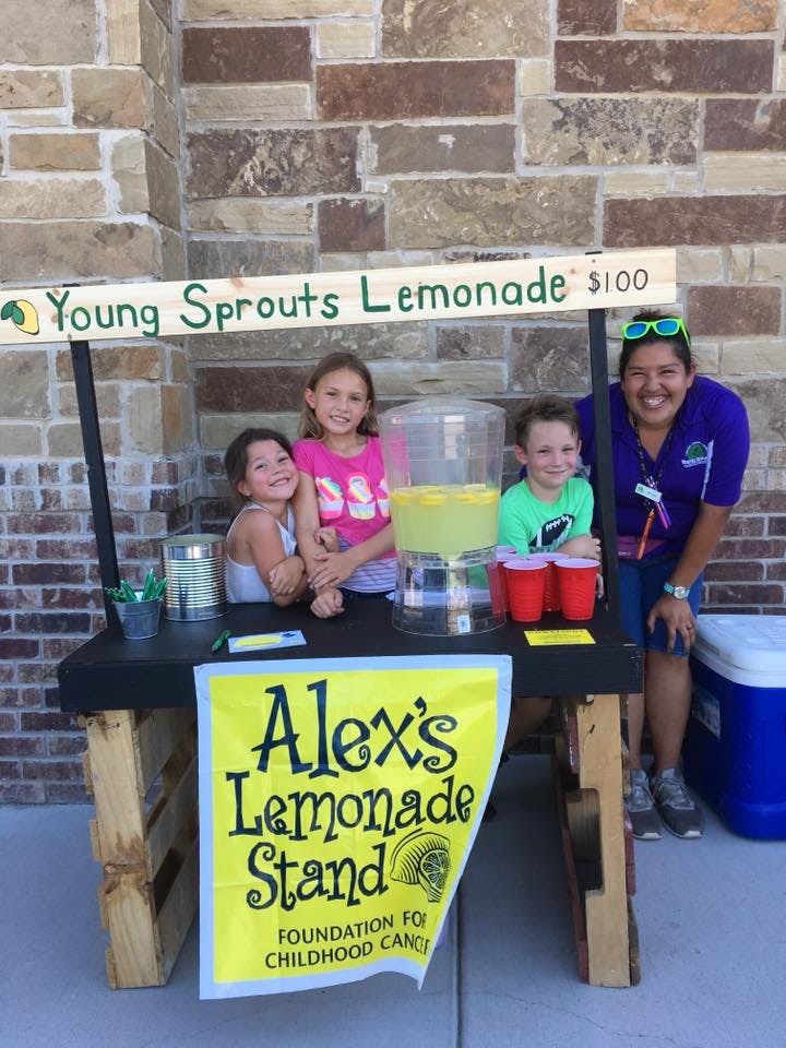 Three children and one adult behind a lemonade stand with a sign that says "Young Sprouts Lemonade"