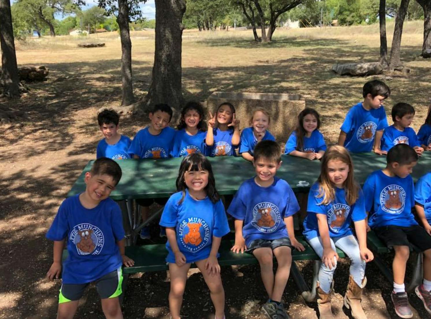 Group of children in blue shirts sitting around a picnic table outdoors
