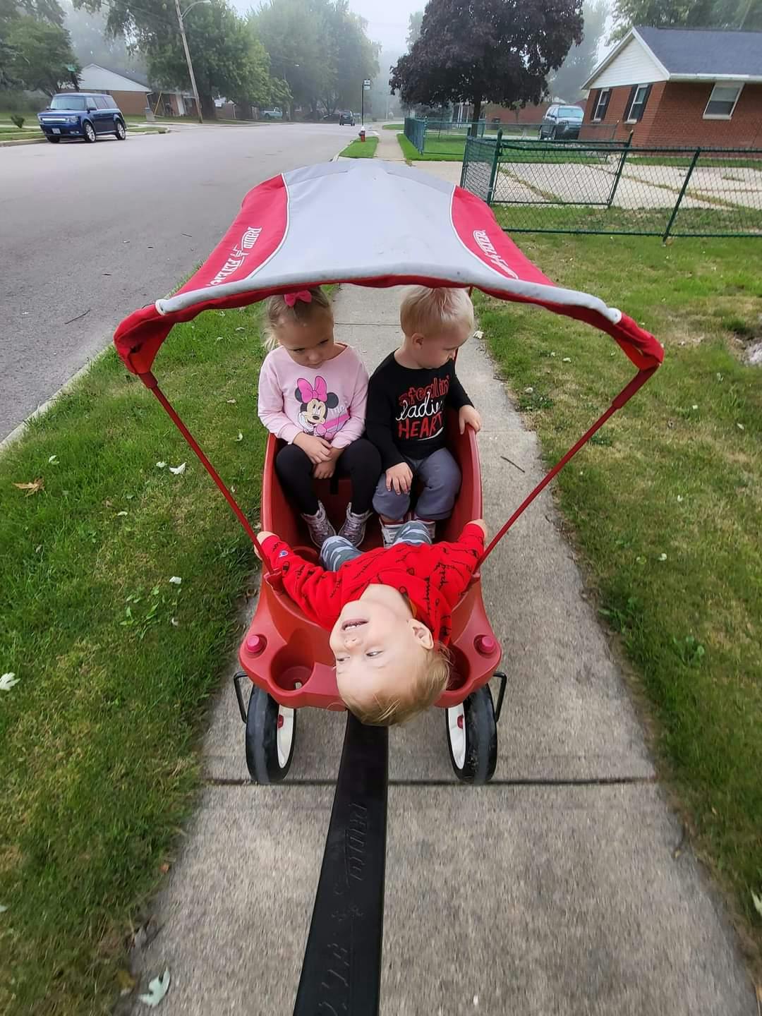 Three children in a red wagon with a canopy, one child hanging out of the wagon