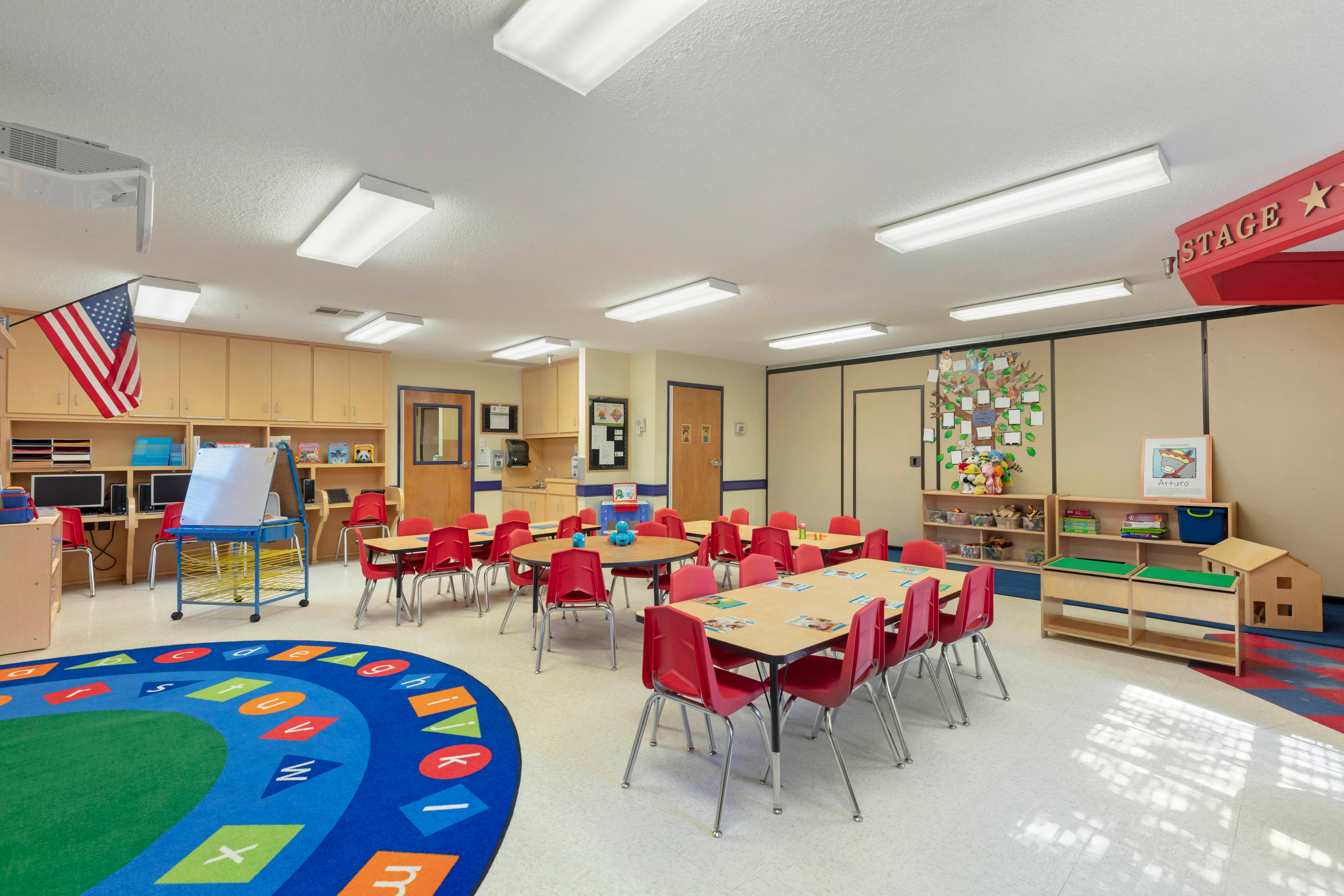Classroom with small tables and red chairs, computer stations, and an alphabet rug