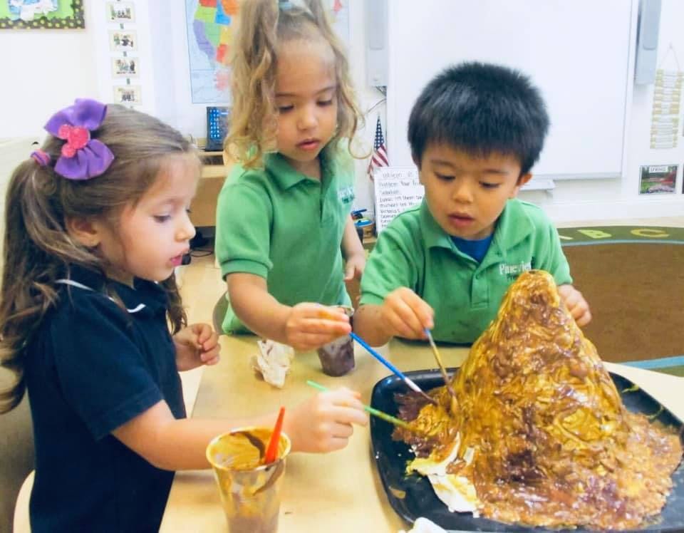 Three children painting a volcano model in a classroom