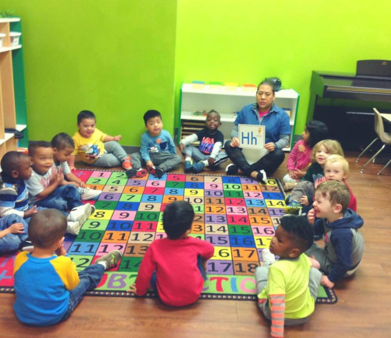 Person holding up a letter H to a group of children sitting on a colorful rug