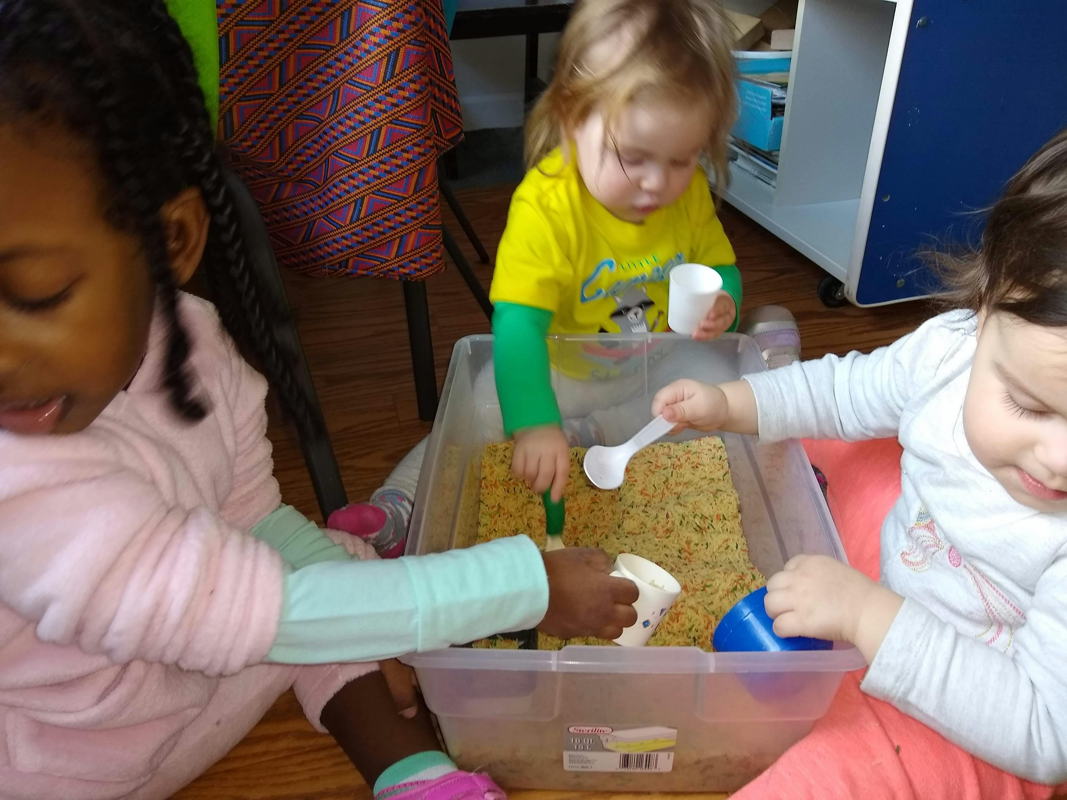 Three children playing with rice and scoops in a clear bin