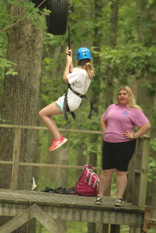 Child on a zip line wearing a helmet and harness, person standing on a platform