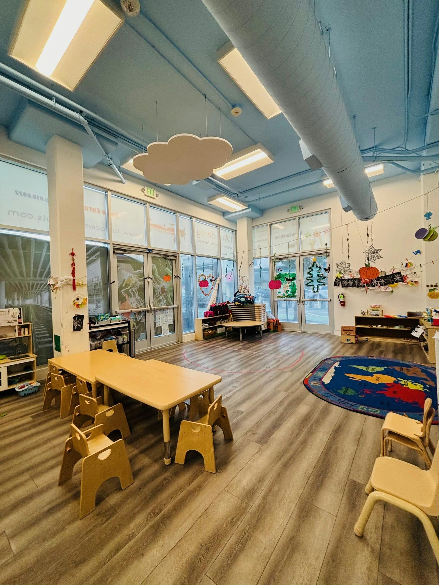 Empty classroom with small tables and chairs, a play rug, and decorated windows